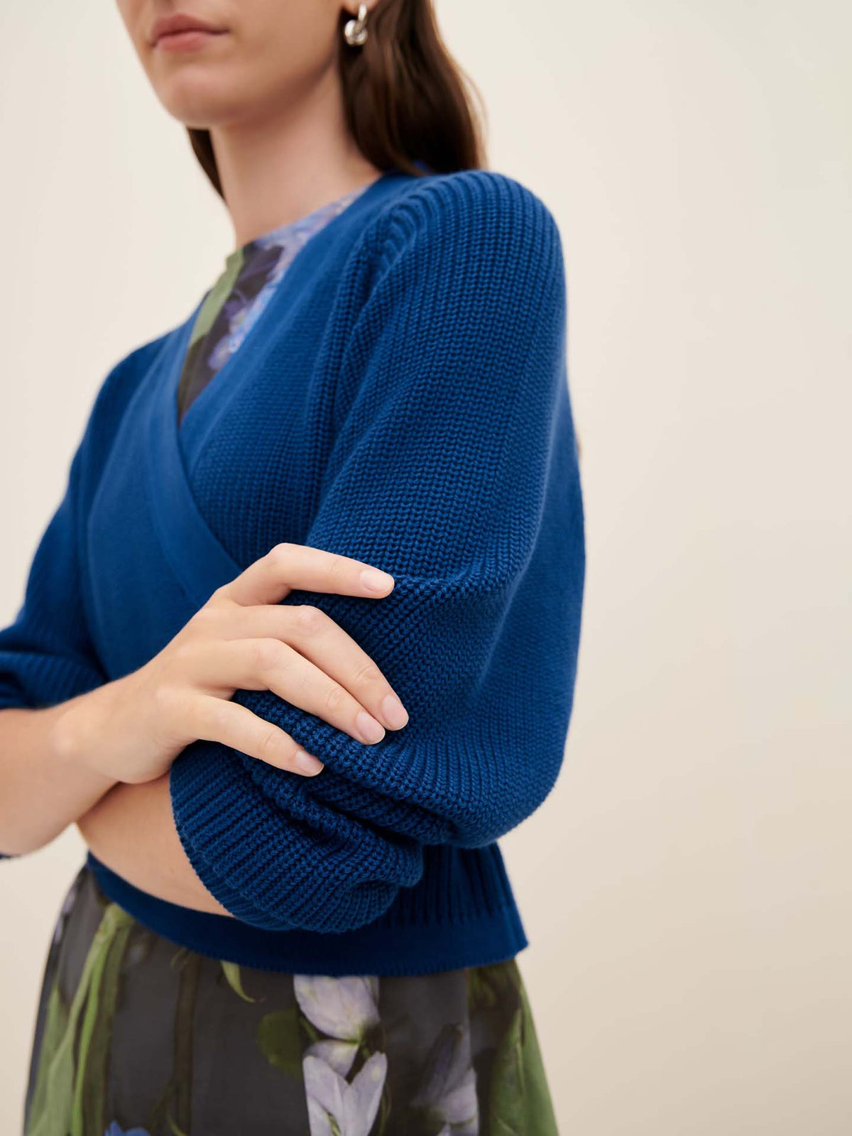 A person wearing the Composure Cardigan in Primary Blue from Kowtow stands with arms crossed, paired with a floral dress.