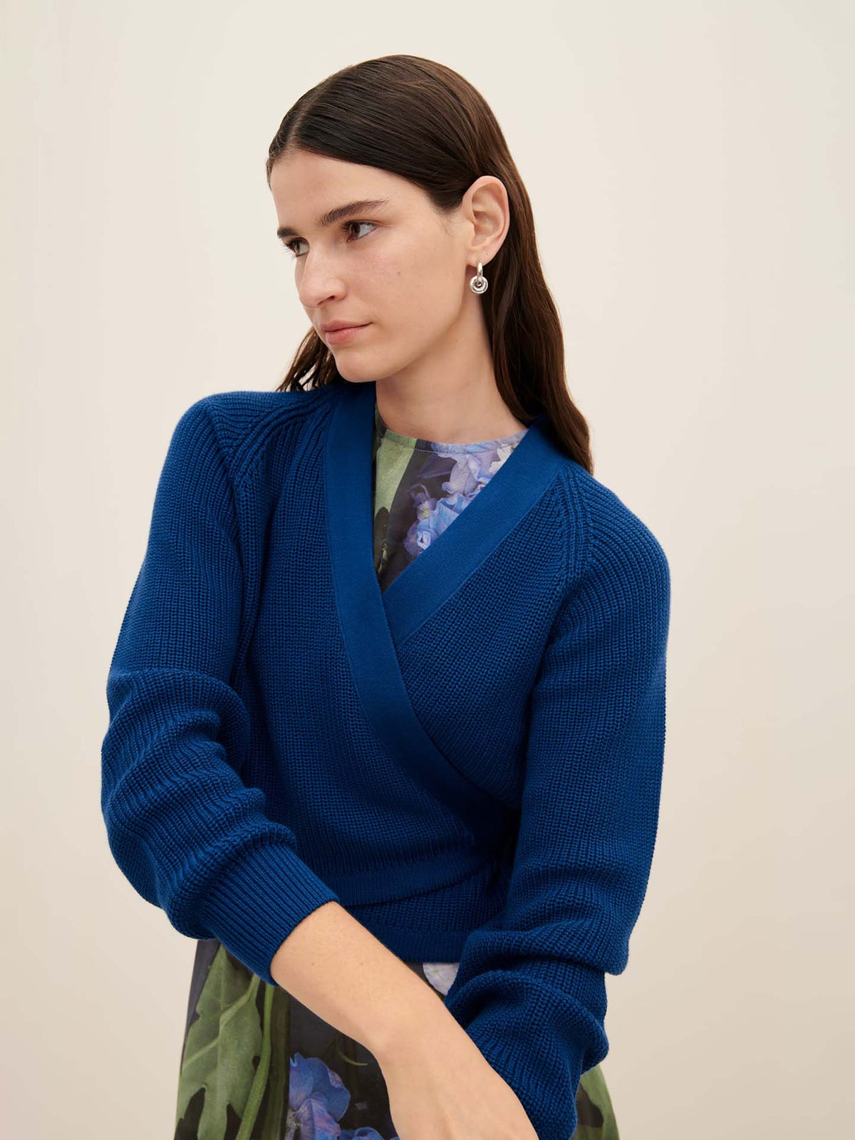 A person with long dark hair wearing an oversized Composure Cardigan in Primary Blue by Kowtow and a floral dress stands against a plain background.