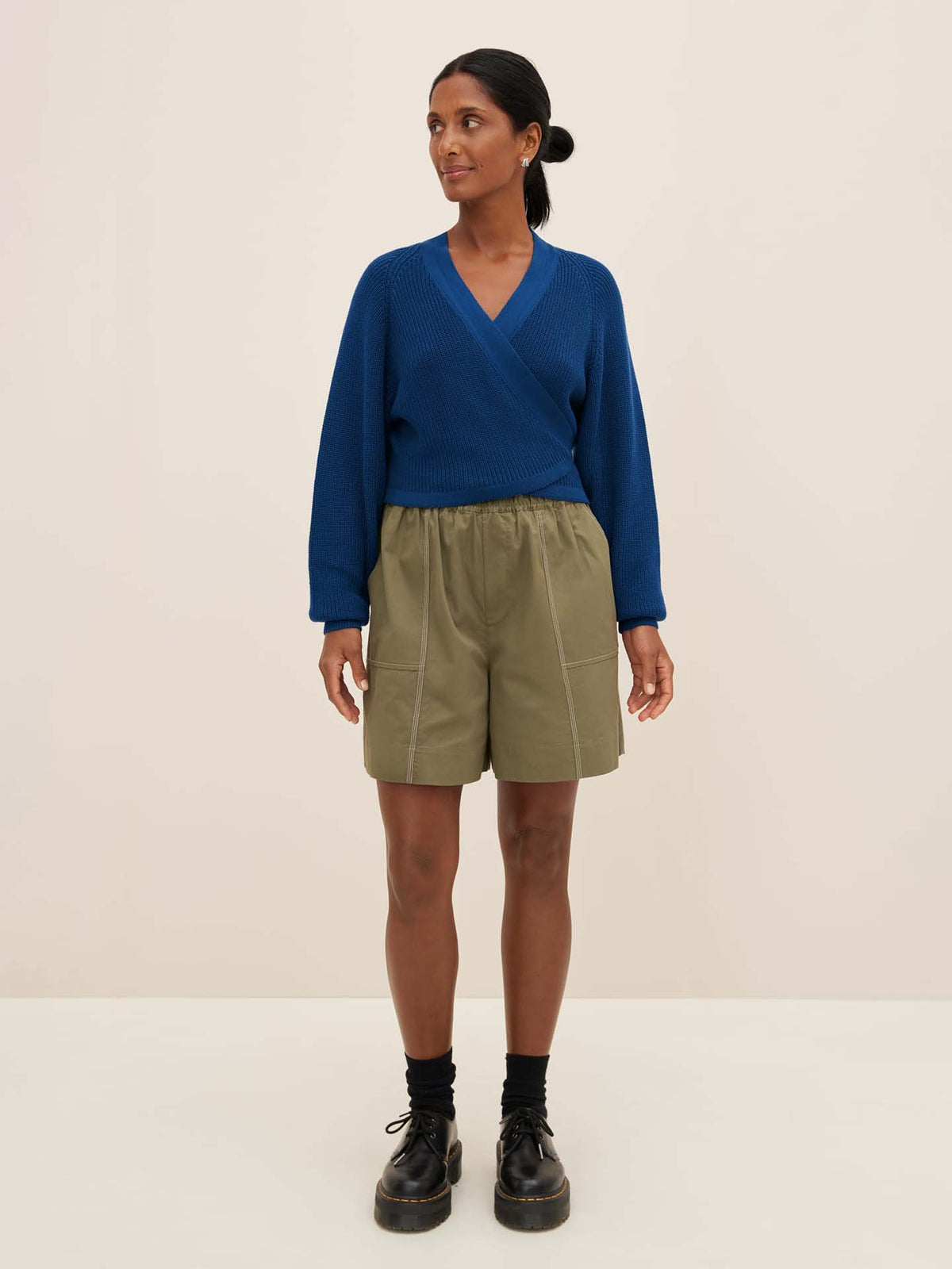 A person wearing the Composure Cardigan in Primary Blue by Kowtow, olive green shorts, and black shoes stands on a plain background.