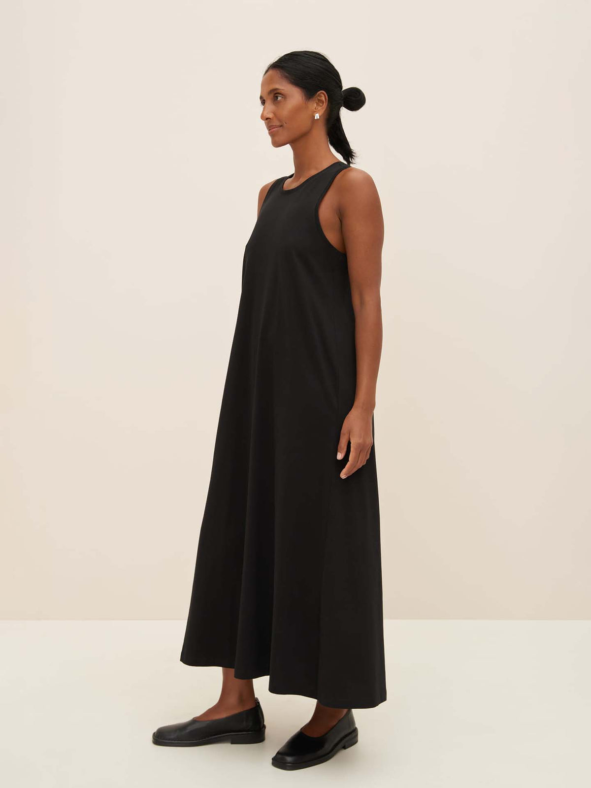 A person in a Kowtow Cross Back Dress – Black, paired with black shoes, stands against a plain, light background, showcasing the standard fit.