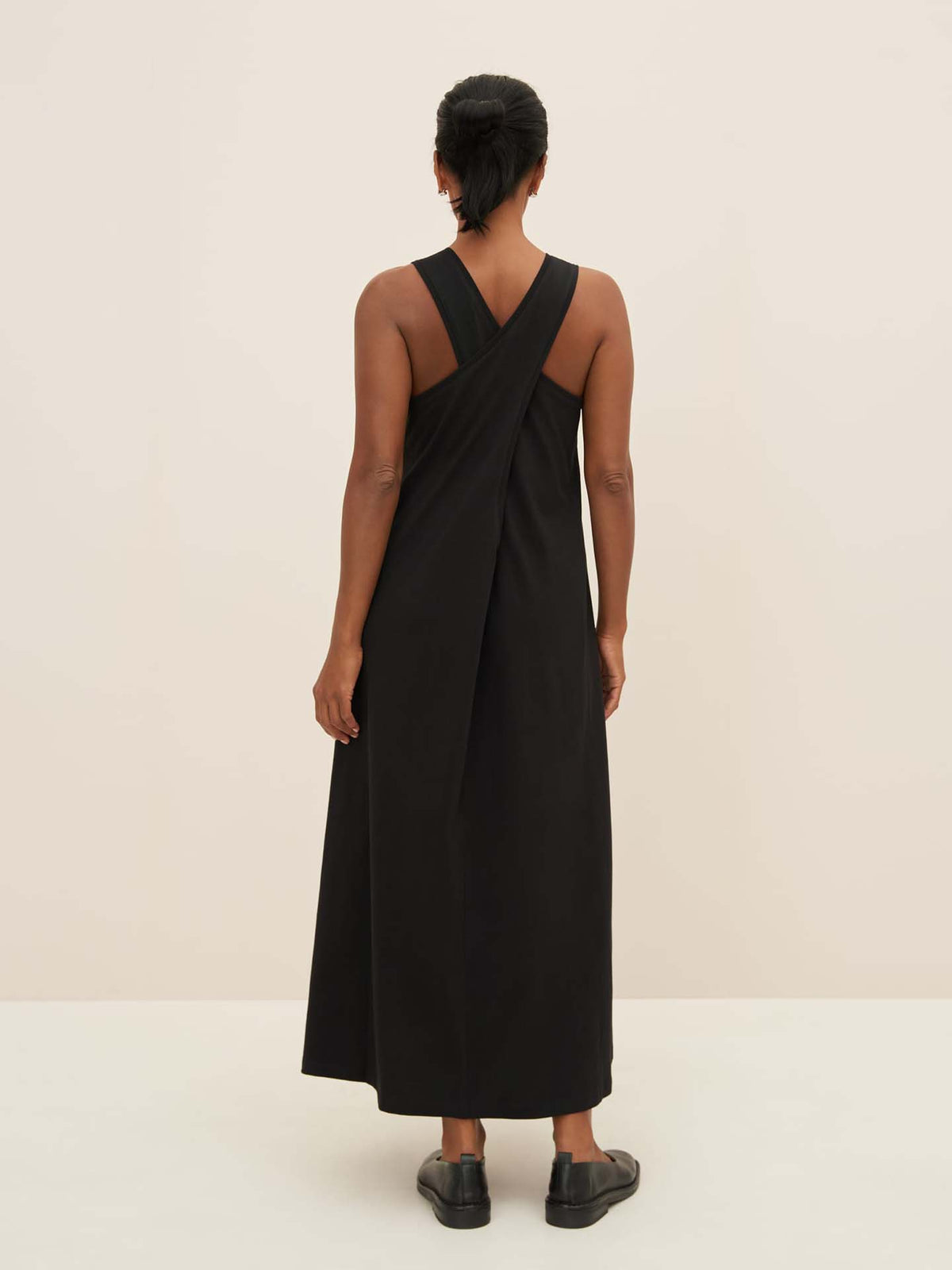 Against a neutral backdrop, an individual stands with their back turned, donning Kowtow&#39;s Cross Back Dress in black, paired with matching shoes to perfectly illustrate the brand&#39;s standard size guide fit.