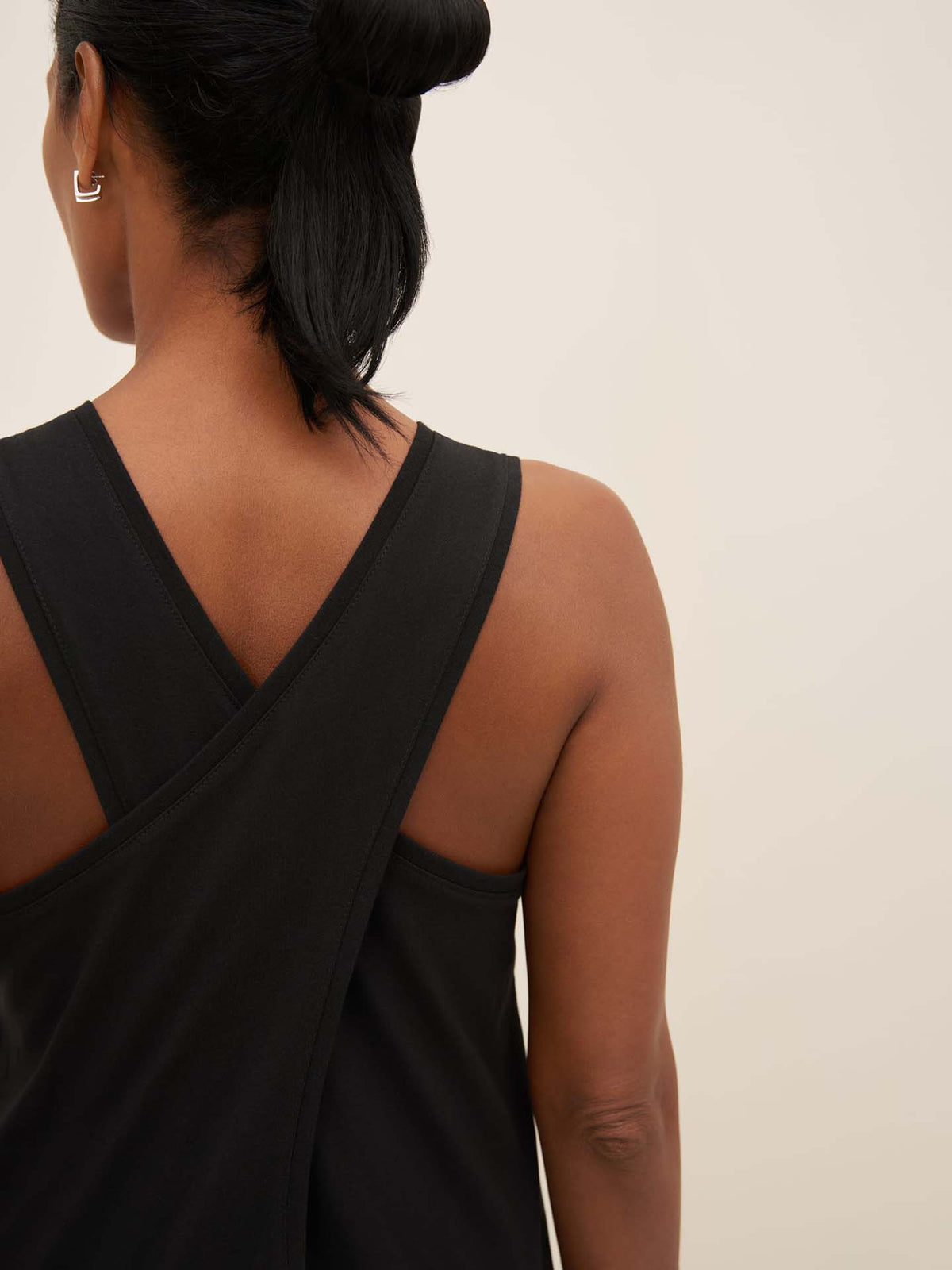 Back view of a person with black hair in a bun, wearing the Kowtow Cross Back Dress in black, standing against a plain background.