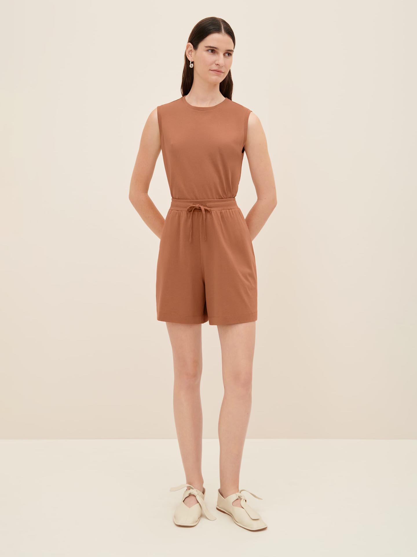 A woman stands against a plain background wearing the Drawcord Shorts in Sienna from Kowtow and cream-colored shoes. She has long hair and is looking directly at the camera, with her arms relaxed by her sides. Consult our size guide for the perfect fit.