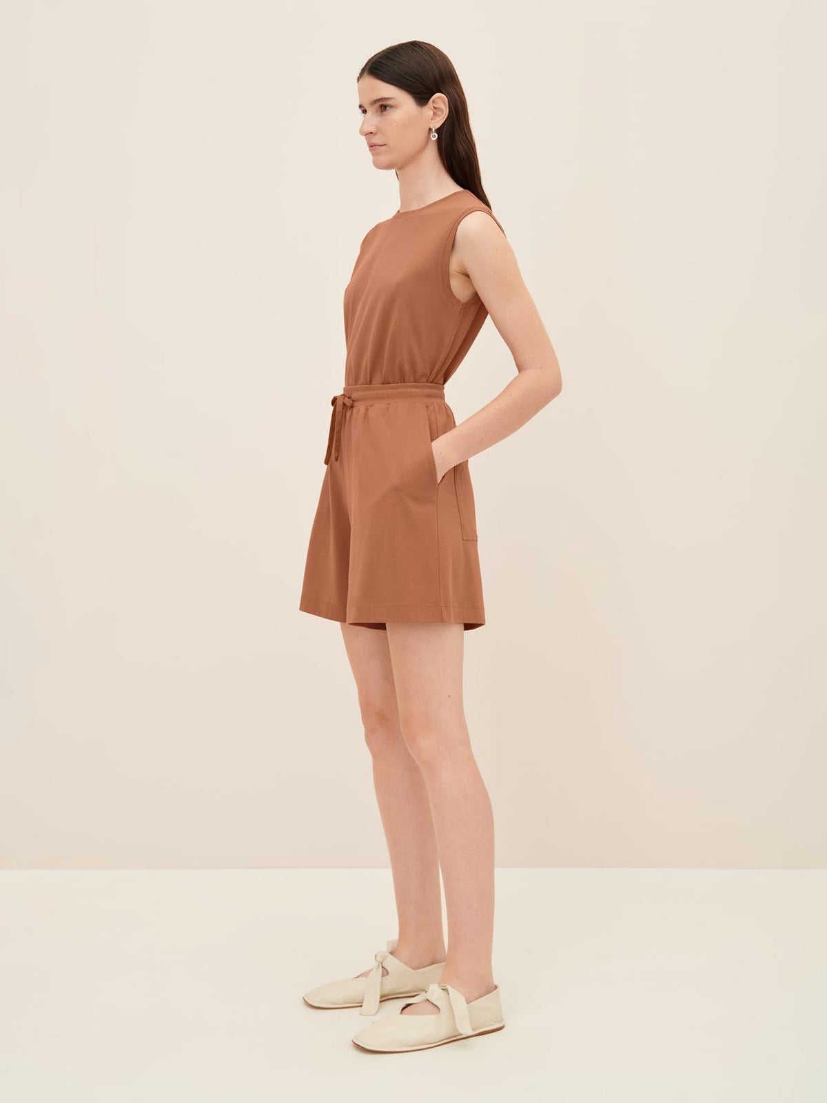 A person stands against a plain background, wearing Kowtow&#39;s relaxed-fit Drawcord Shorts in sienna with a tie at the waist and beige shoes. The outfit is effortlessly chic.