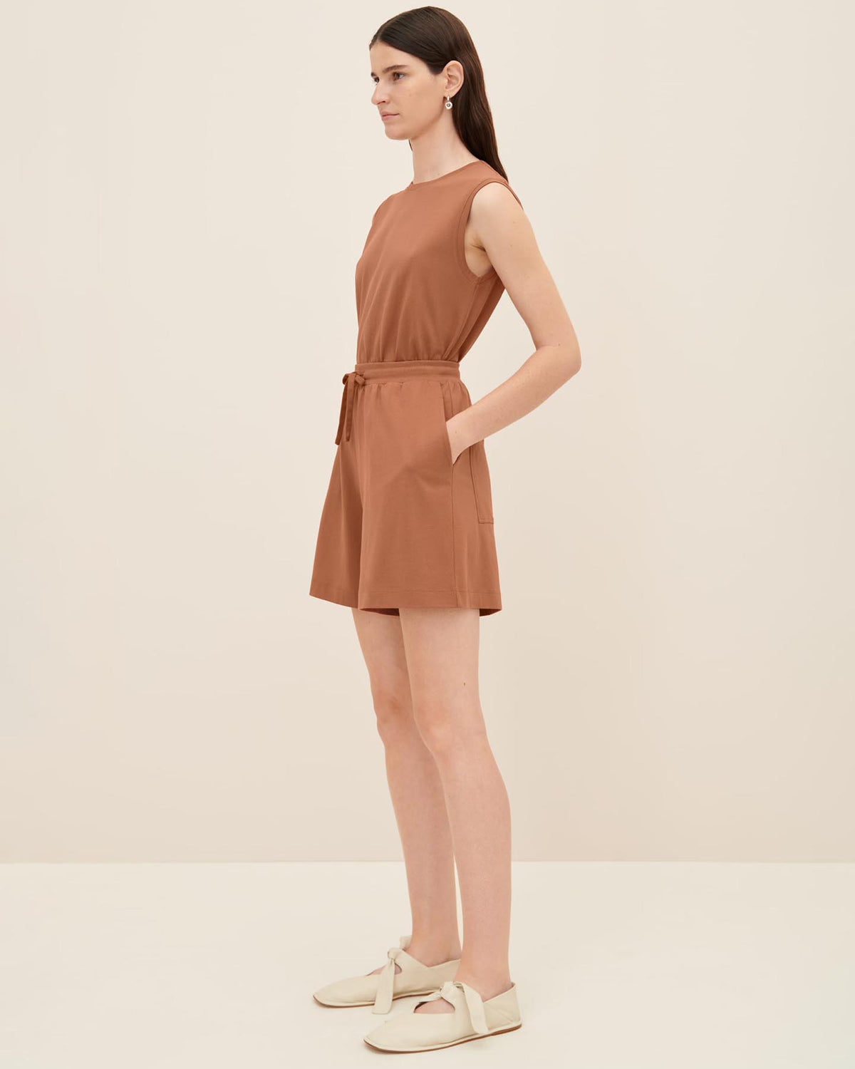 A person stands against a plain background, wearing Kowtow's relaxed-fit Drawcord Shorts in sienna with a tie at the waist and beige shoes. The outfit is effortlessly chic.