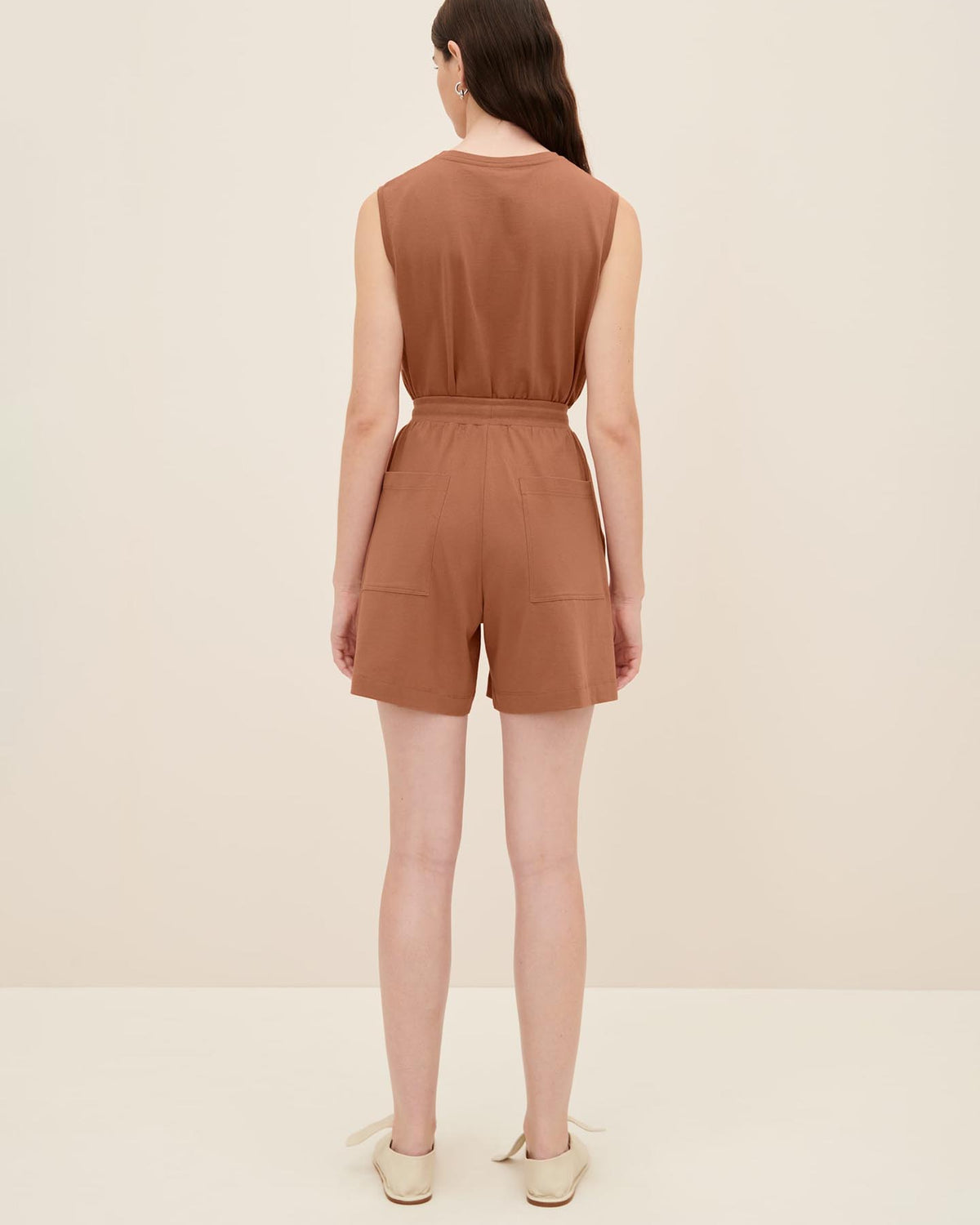 A person with long hair is standing and facing away, wearing a sleeveless brown top and Kowtow's Drawcord Shorts in Sienna, featuring a relaxed fit, in a well-lit indoor setting.