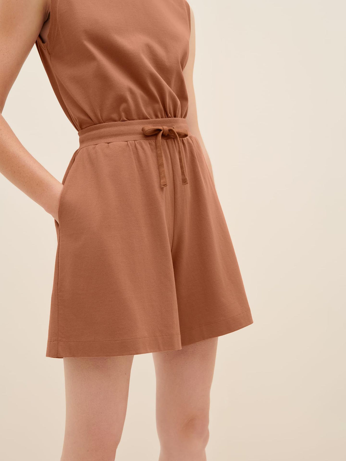 A person wearing a sleeveless romper in the color Sienna, with their hands in the pockets of Kowtow&#39;s Drawcord Shorts, showcasing the drawstring waist design for a relaxed fit.