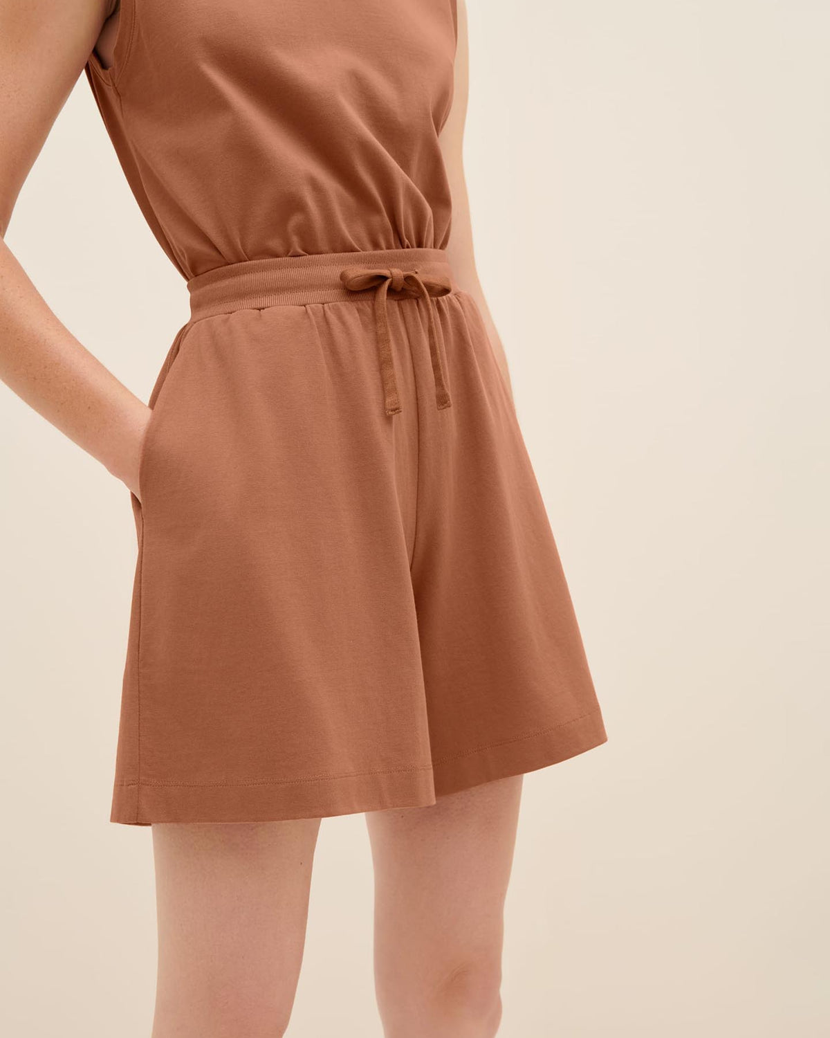 A person wearing a sleeveless romper in the color Sienna, with their hands in the pockets of Kowtow's Drawcord Shorts, showcasing the drawstring waist design for a relaxed fit.
