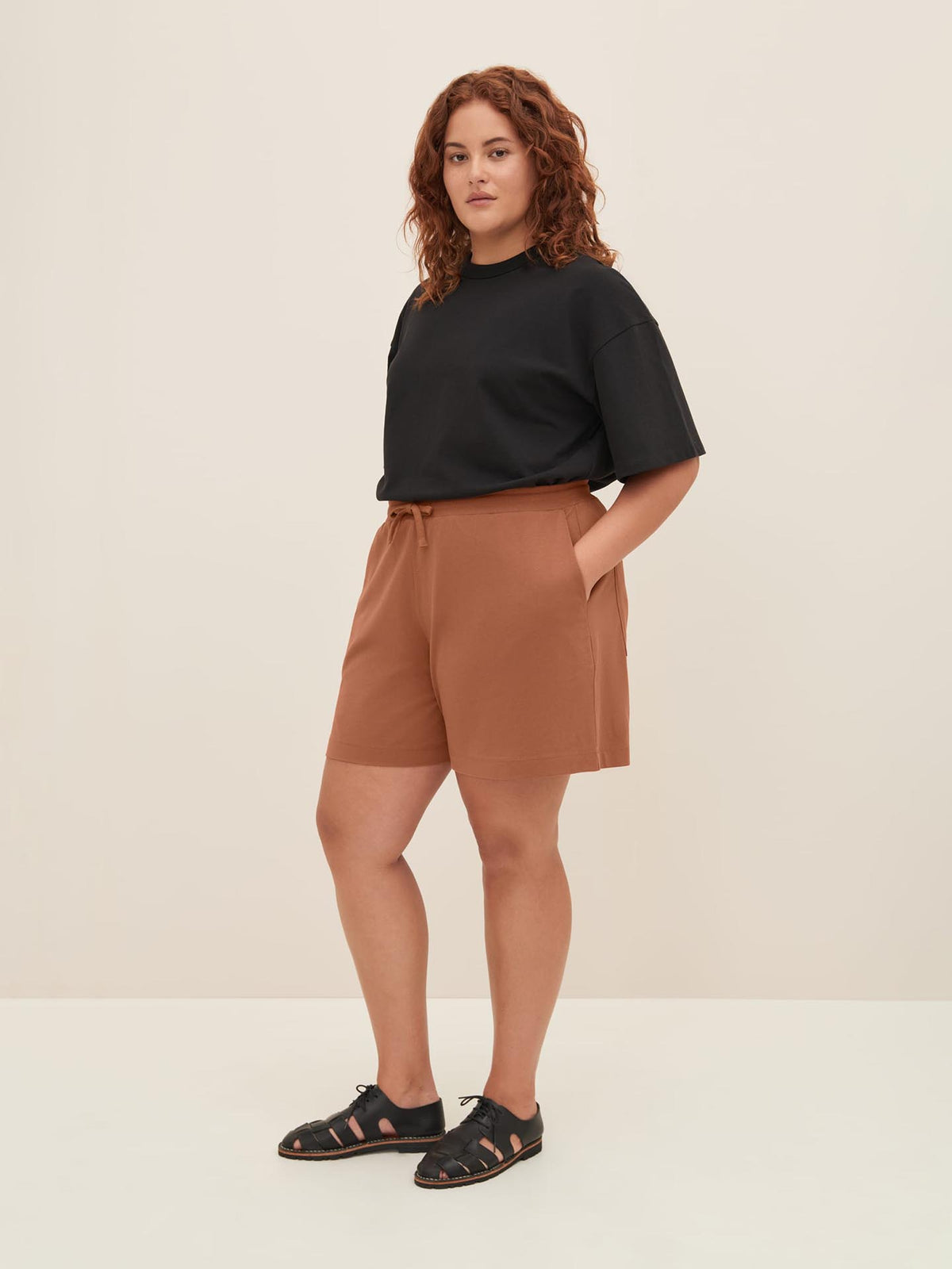 A person with curly hair wearing a black t-shirt, Kowtow&#39;s Drawcord Shorts in Sienna, and black sandals stands facing the camera with hands in pockets against a plain background. The relaxed fit of the outfit offers comfort without compromising style.