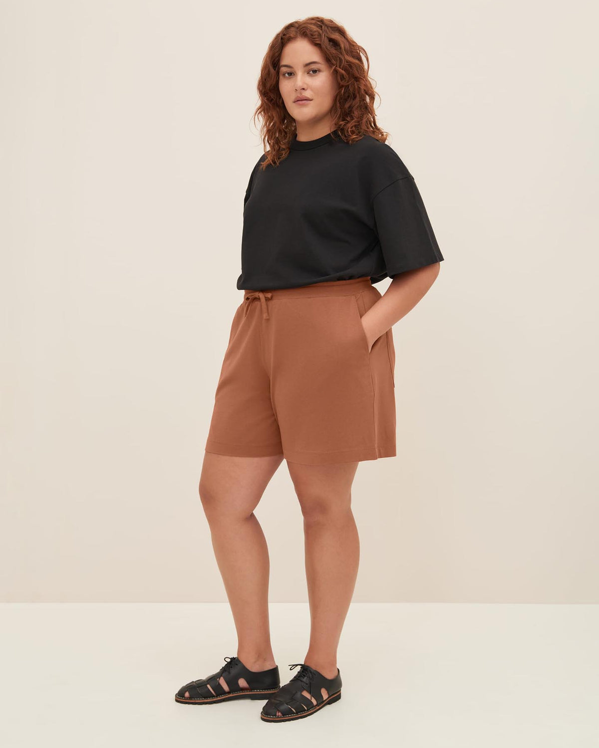 A person with curly hair wearing a black t-shirt, Kowtow's Drawcord Shorts in Sienna, and black sandals stands facing the camera with hands in pockets against a plain background. The relaxed fit of the outfit offers comfort without compromising style.