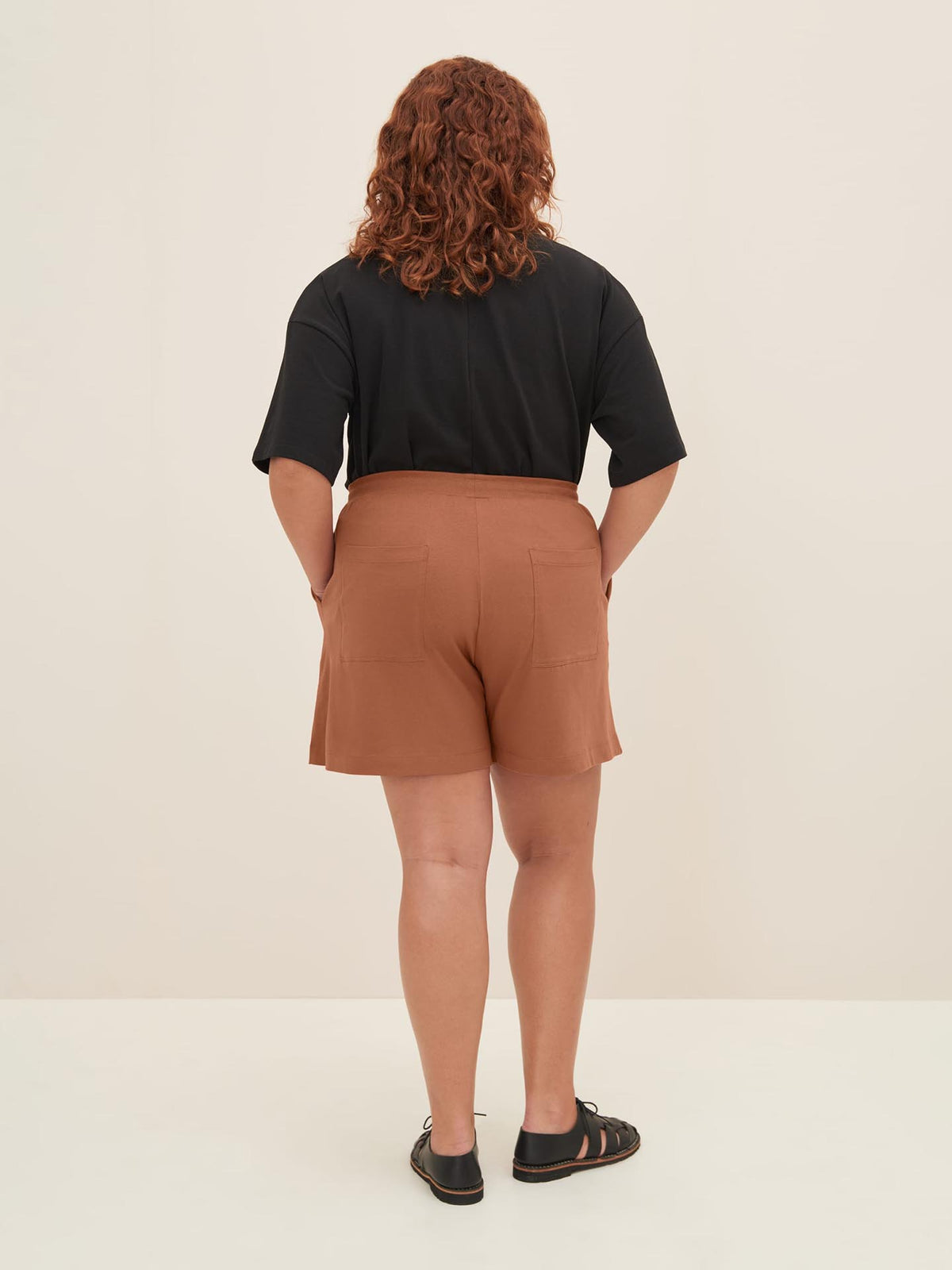 A person with curly hair stands facing away, wearing a black top, Kowtow&#39;s Drawcord Shorts in Sienna with a relaxed fit at the natural waist, and black sandals.