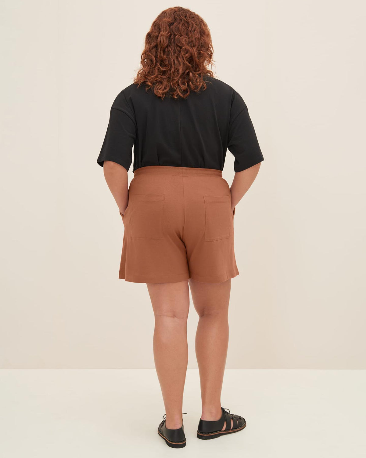 A person with curly hair stands facing away, wearing a black top, Kowtow's Drawcord Shorts in Sienna with a relaxed fit at the natural waist, and black sandals.