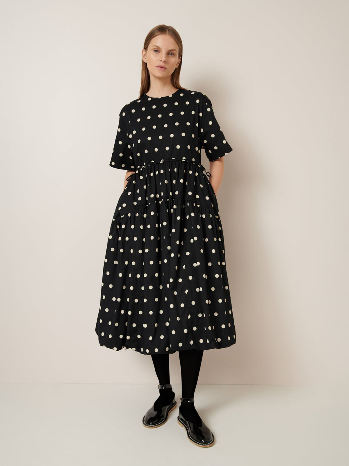 A person stands against a plain backdrop wearing the Eclipse Dress - Polka Dot by Kowtow, paired with relaxed fit leggings and black shoes.