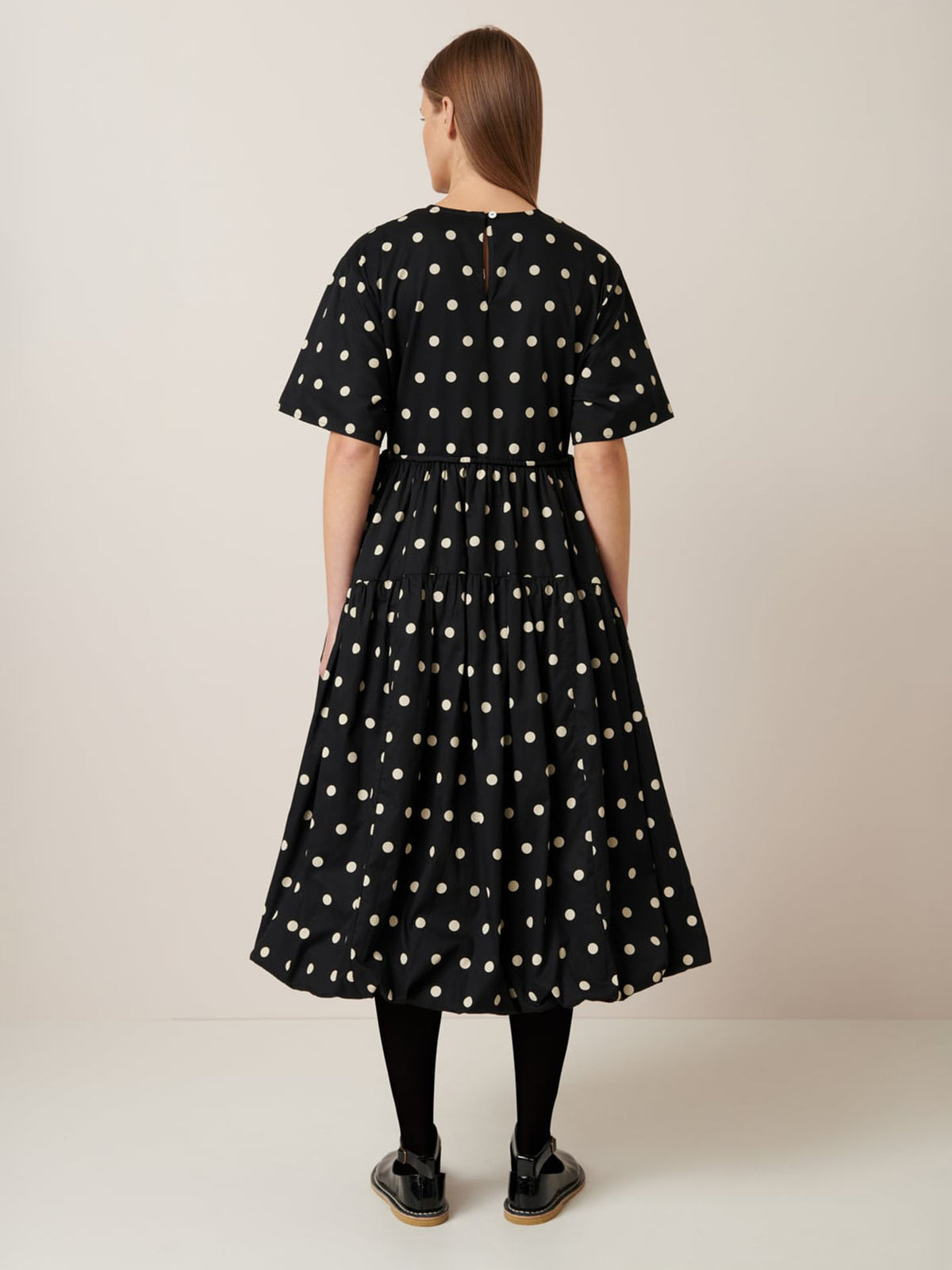 Person facing away in a Kowtow Eclipse Dress – Polka Dot, showcasing short sleeves and a flared skirt for a relaxed fit. Paired with black tights and sandals against a plain backdrop. Refer to the size guide for accurate garment measurements.