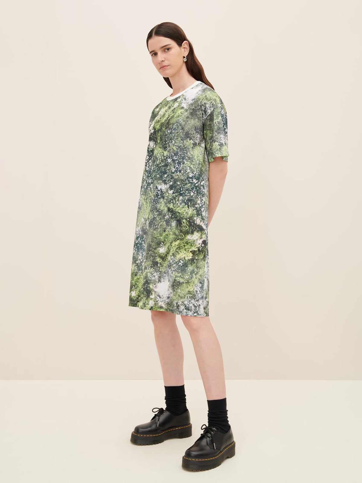 A person stands against a neutral background wearing the Kowtow Foliage Dress, accompanied by black socks and black shoes, all in standard fit.