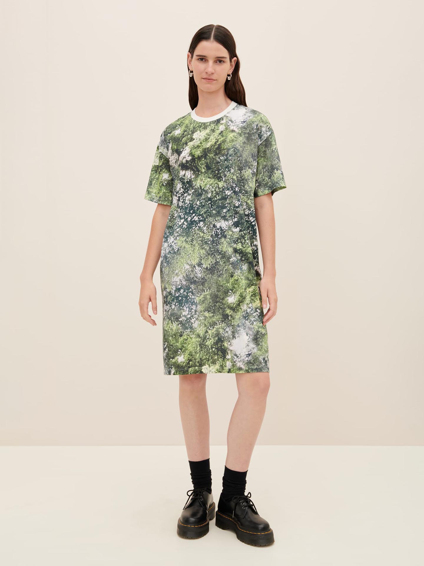 A person standing against a plain background is wearing the Kowtow Foliage Dress, which features a green and white pattern and offers a standard fit, paired with black socks and black shoes.