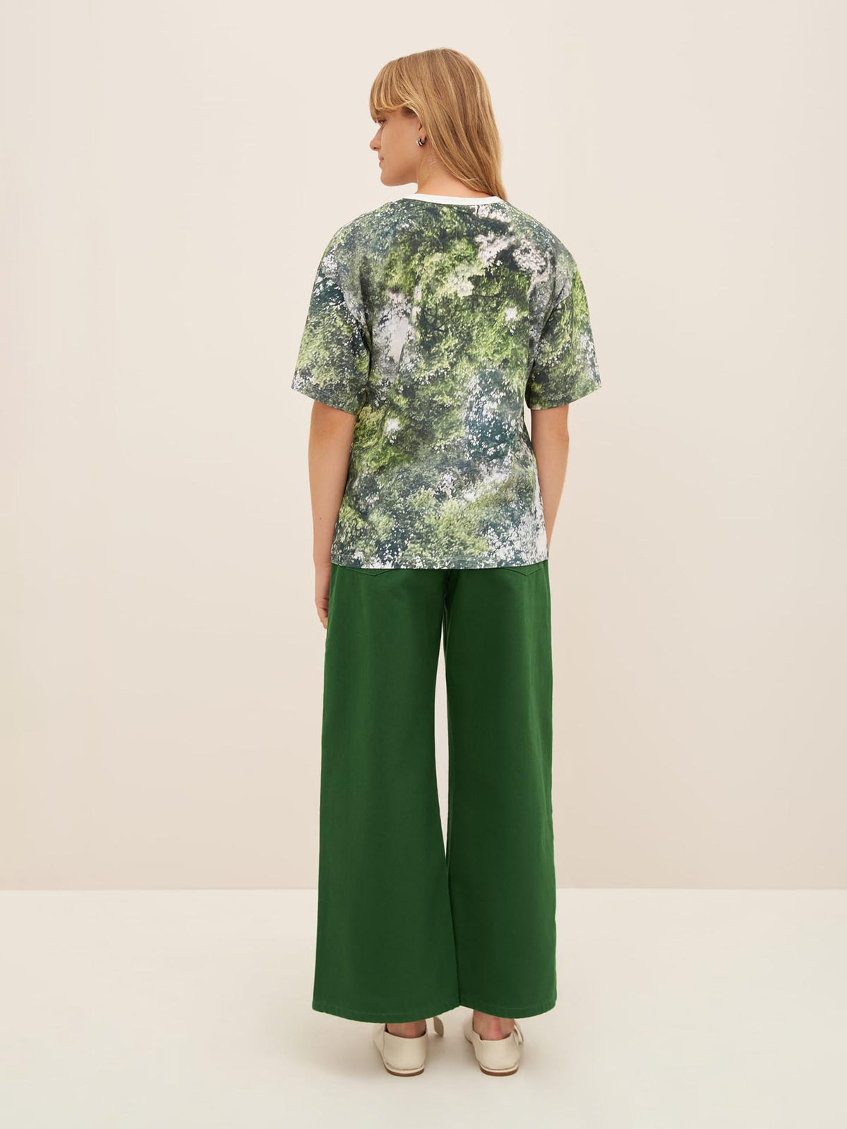 A person with blonde hair stands facing away, wearing an oversized Foliage Tee by Kowtow, green wide-leg pants, and light-colored shoes.