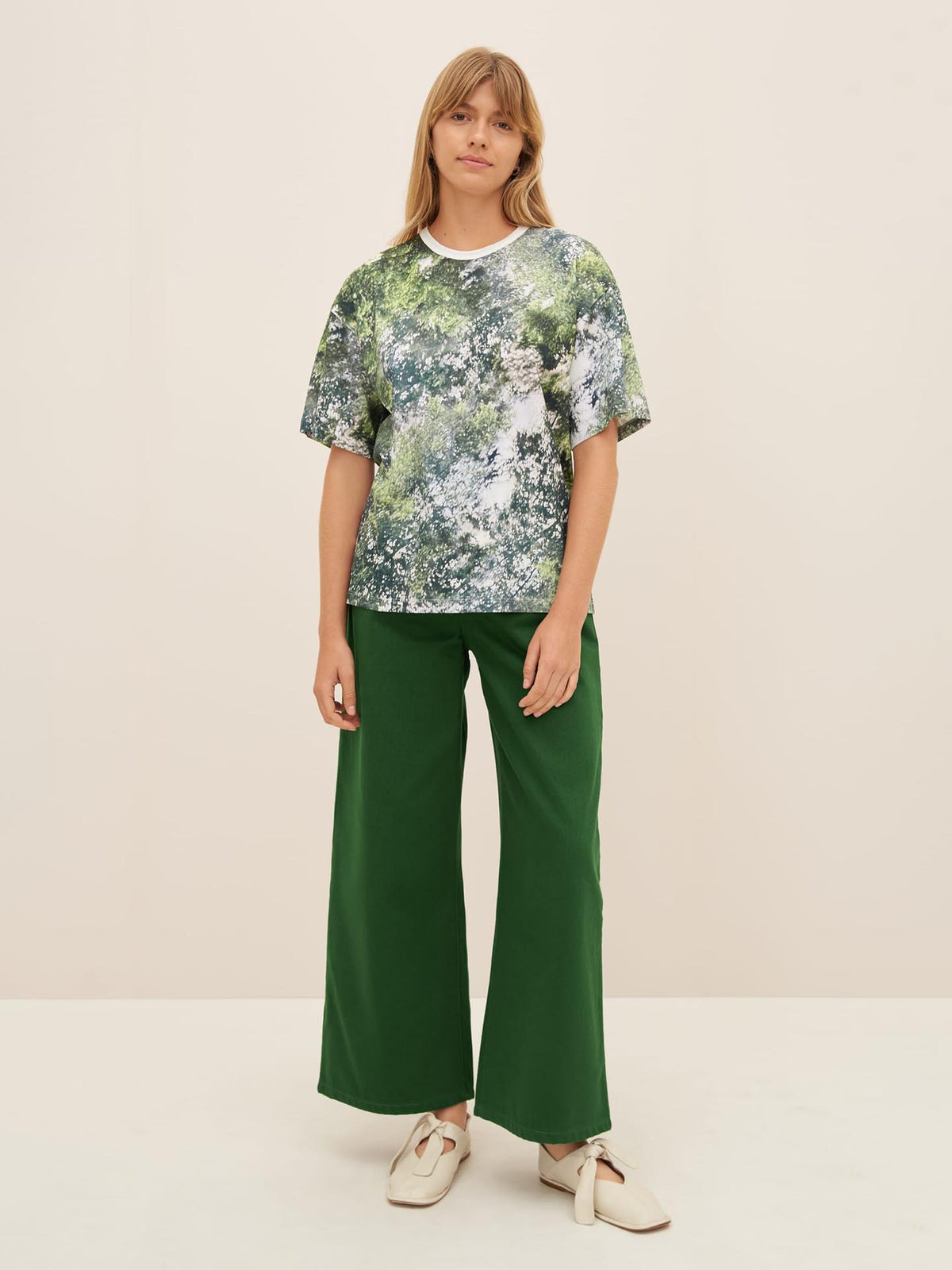 A person wearing the Kowtow Foliage Tee, featuring a green and white pattern with an oversized fit, paired with green wide-leg pants and white shoes, stands against a beige background.