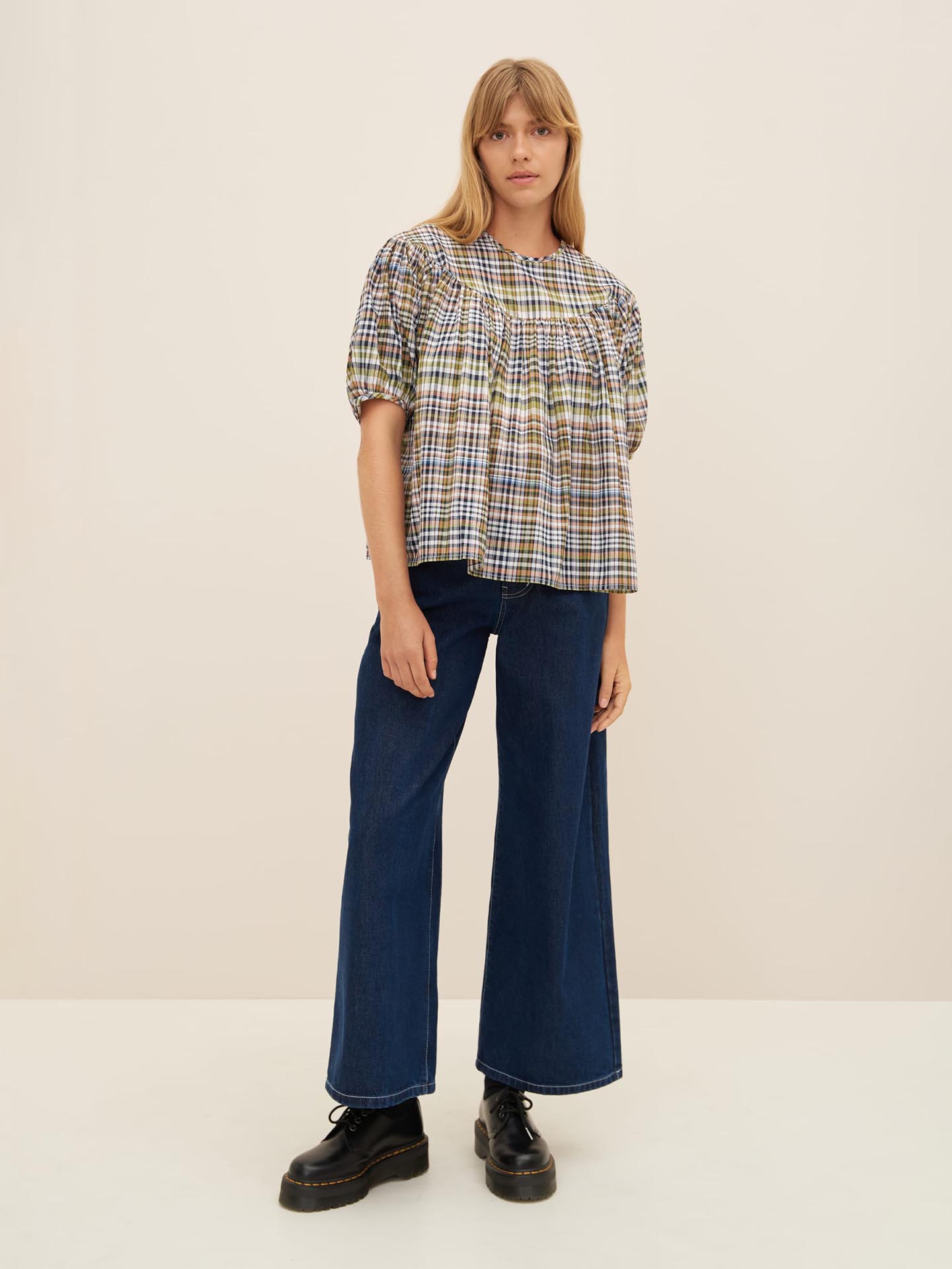 Woman stands against a plain background wearing an oversized fit Frances Top by Kowtow, wide-leg blue jeans, and black platform shoes. She has long blonde hair and a neutral expression. For best results, refer to the size guide when considering sizing down for a perfect fit.