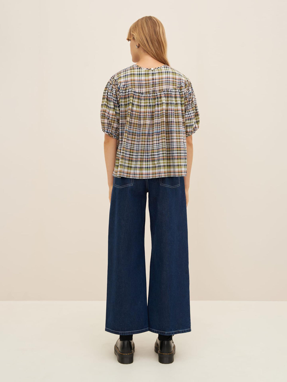 A person with long hair is standing facing away, wearing the Frances Top by Kowtow, which features a checkered pattern and an oversized fit, along with wide-leg denim pants and black shoes.