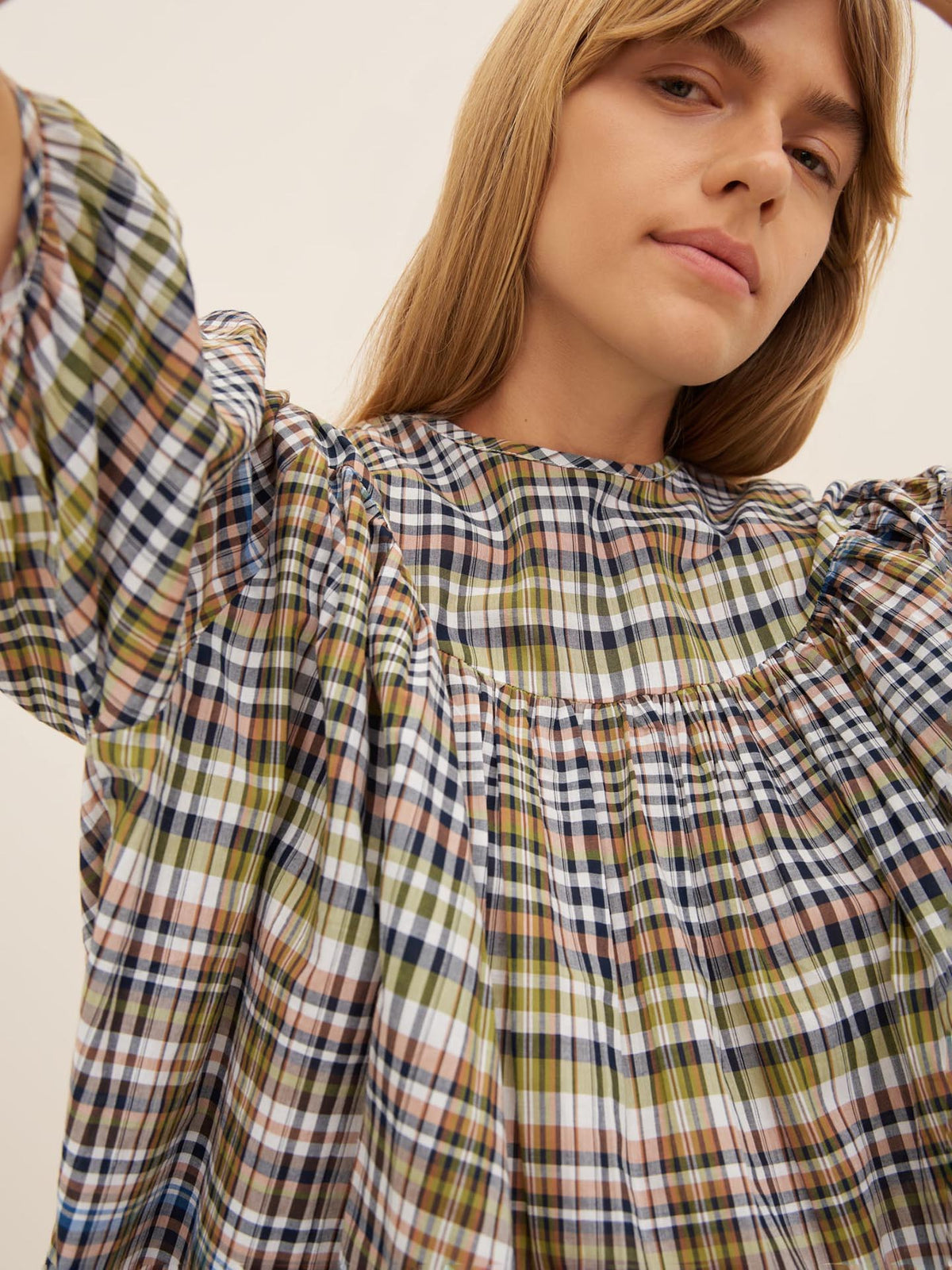 Person wearing the Frances Top by Kowtow in an oversized plaid design with smocked detailing, gazes forward. Their raised arm partially extends out of the frame against a plain, neutral background.