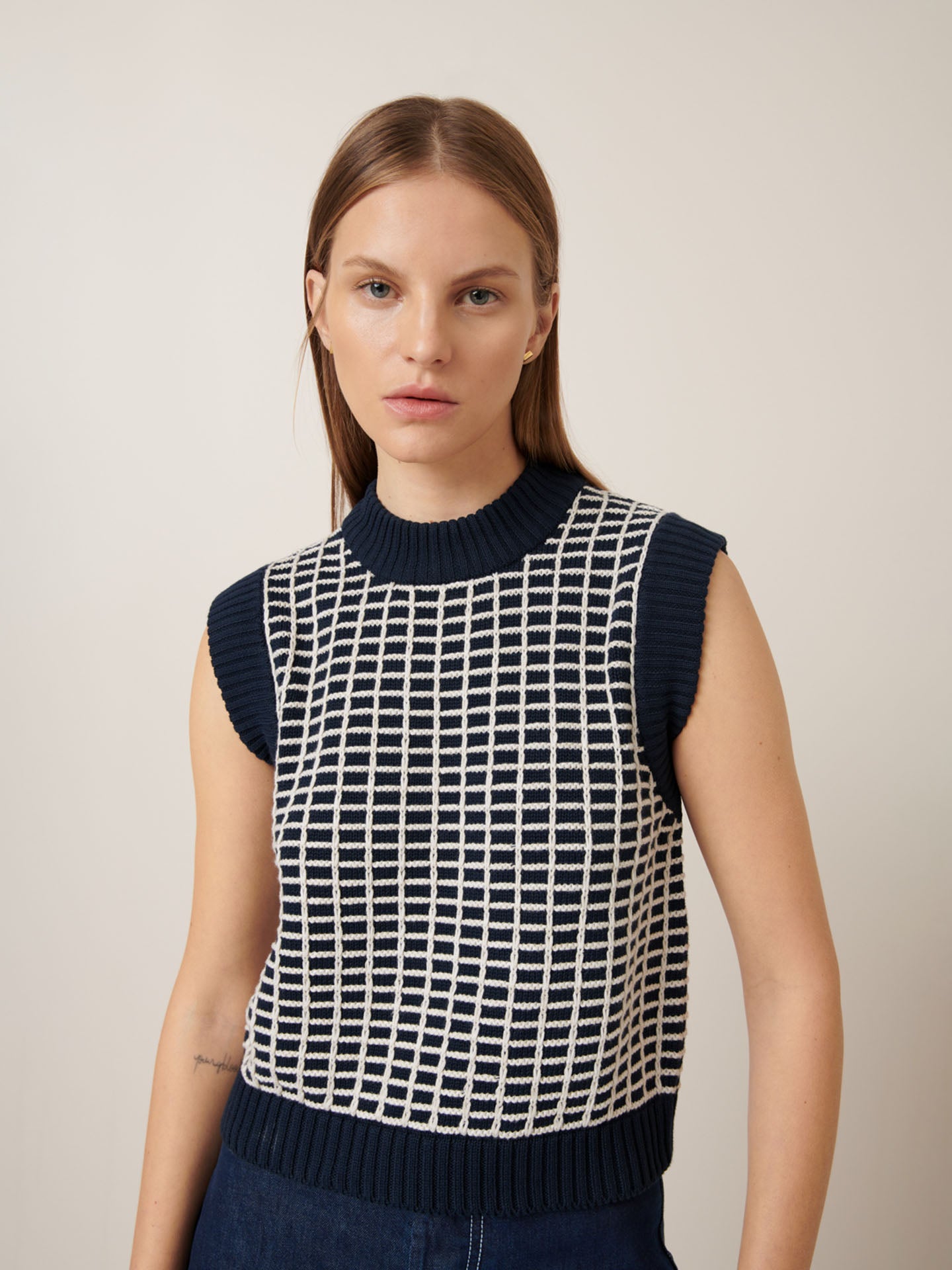 A person with long hair wears the Kowtow Grid Vest in a navy grid pattern, featuring a sleeveless design and relaxed fit, standing against a neutral background.
