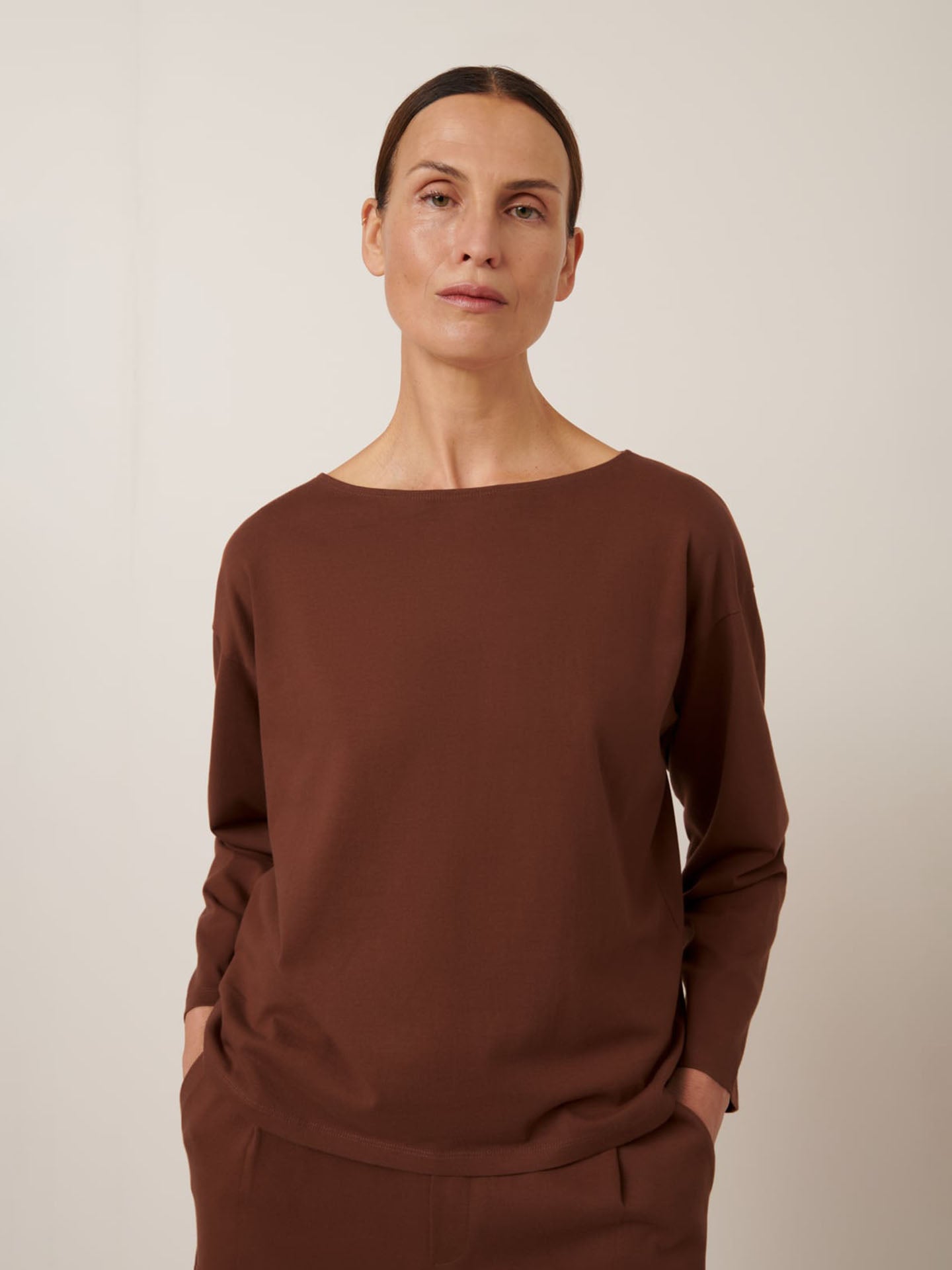 A person with brown hair stands in a relaxed fit, wearing the Jamie Top in Sequoia from Kowtow, paired with brown pants and hands in pockets against a plain backdrop.