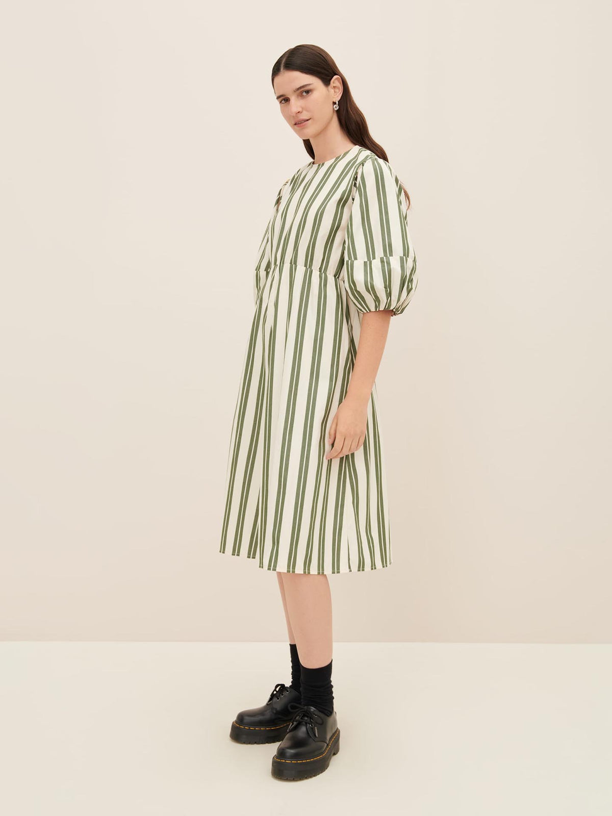 A person stands against a plain background wearing the Joan Dress by Kowtow, featuring white fabric with vertical green stripes and puffy sleeves. They pair it with black shoes and socks, perfectly showcasing the dress in size XS standard fit.