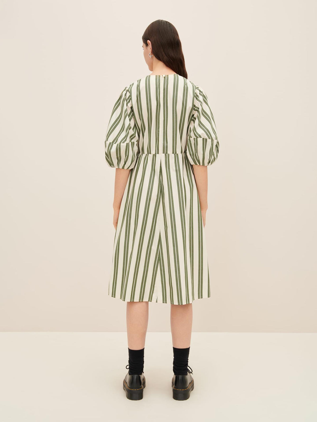 A person stands facing away, wearing a Kowtow Joan Dress in size XS, which is white with vertical green stripes and features puffy sleeves. They have long hair and are wearing black shoes and socks.