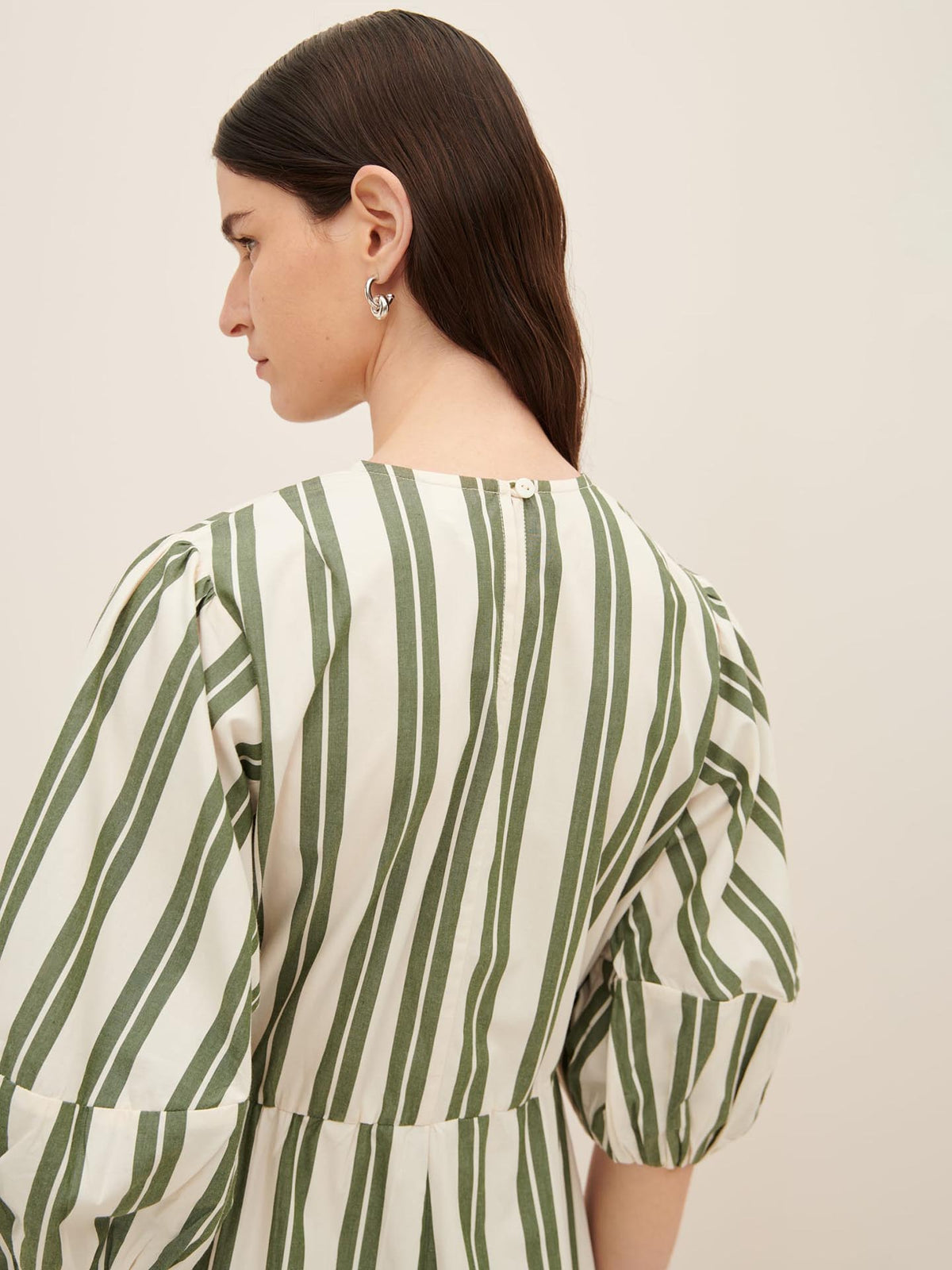 A woman with long dark hair is seen from behind, wearing a Kowtow Joan Dress in size XS, featuring vertical green stripes and balloon sleeves, following the standard fit guide.