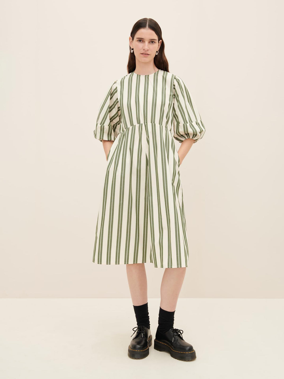 A person stands against a neutral background, wearing a Kowtow Joan Dress in size XS. The white dress features green vertical stripes and has pockets where their hands rest comfortably, paired with black shoes.