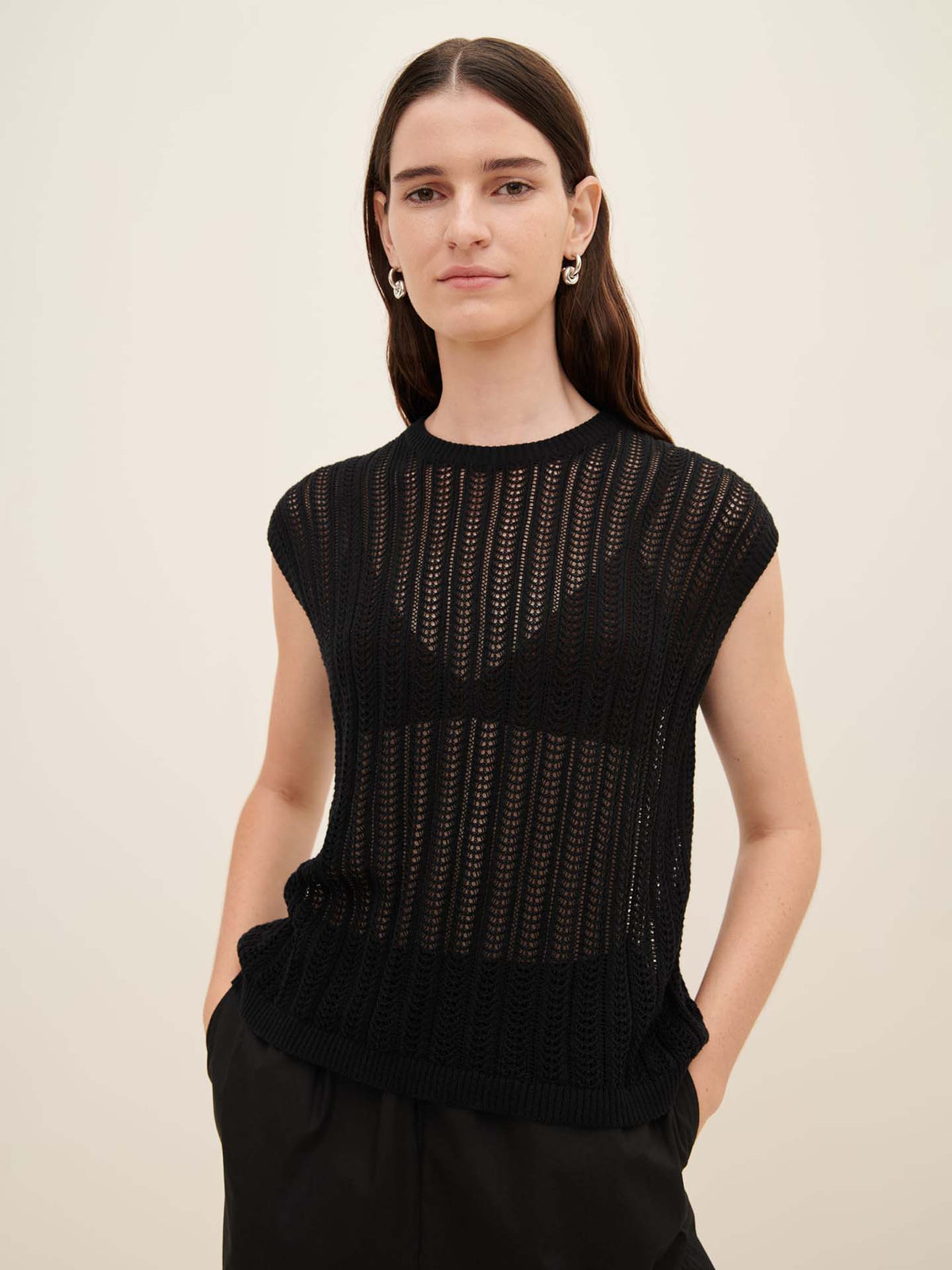 A woman wearing the Kowtow Komorebi Singlet paired with black pants, highlighting the standard fit of Kowtow&#39;s collection, stands with her hands in her pockets against a plain background.