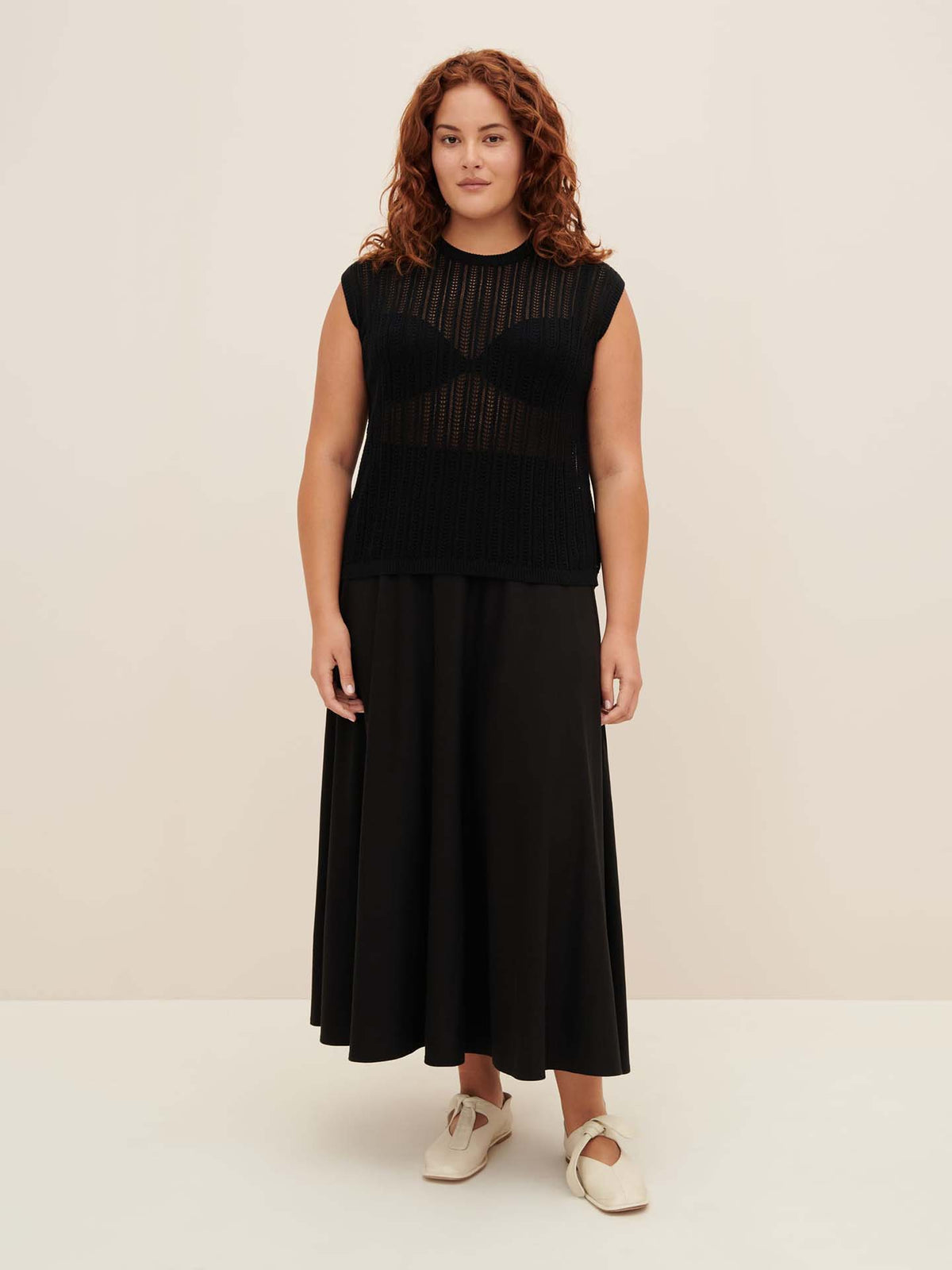 A person with curly red hair is modeling the Komorebi Singlet by Kowtow, paired with a black skirt to highlight its standard fit, set against a plain beige background.