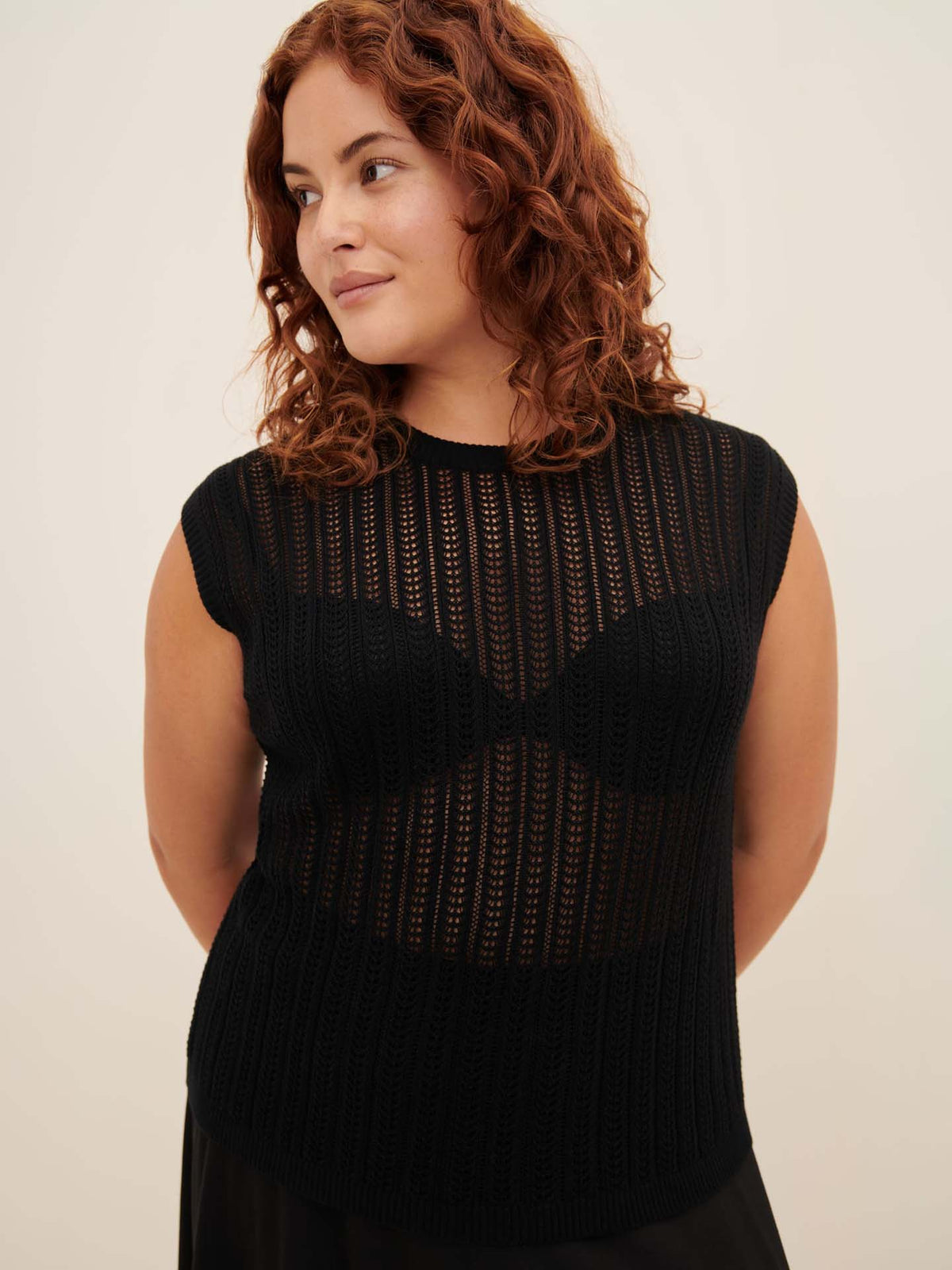 A woman with curly hair stands against a plain background, wearing the Komorebi Singlet from Kowtow—a standard fit, black sleeveless see-through knit top over a black bra. For the perfect fit, refer to the Kowtow size guide.