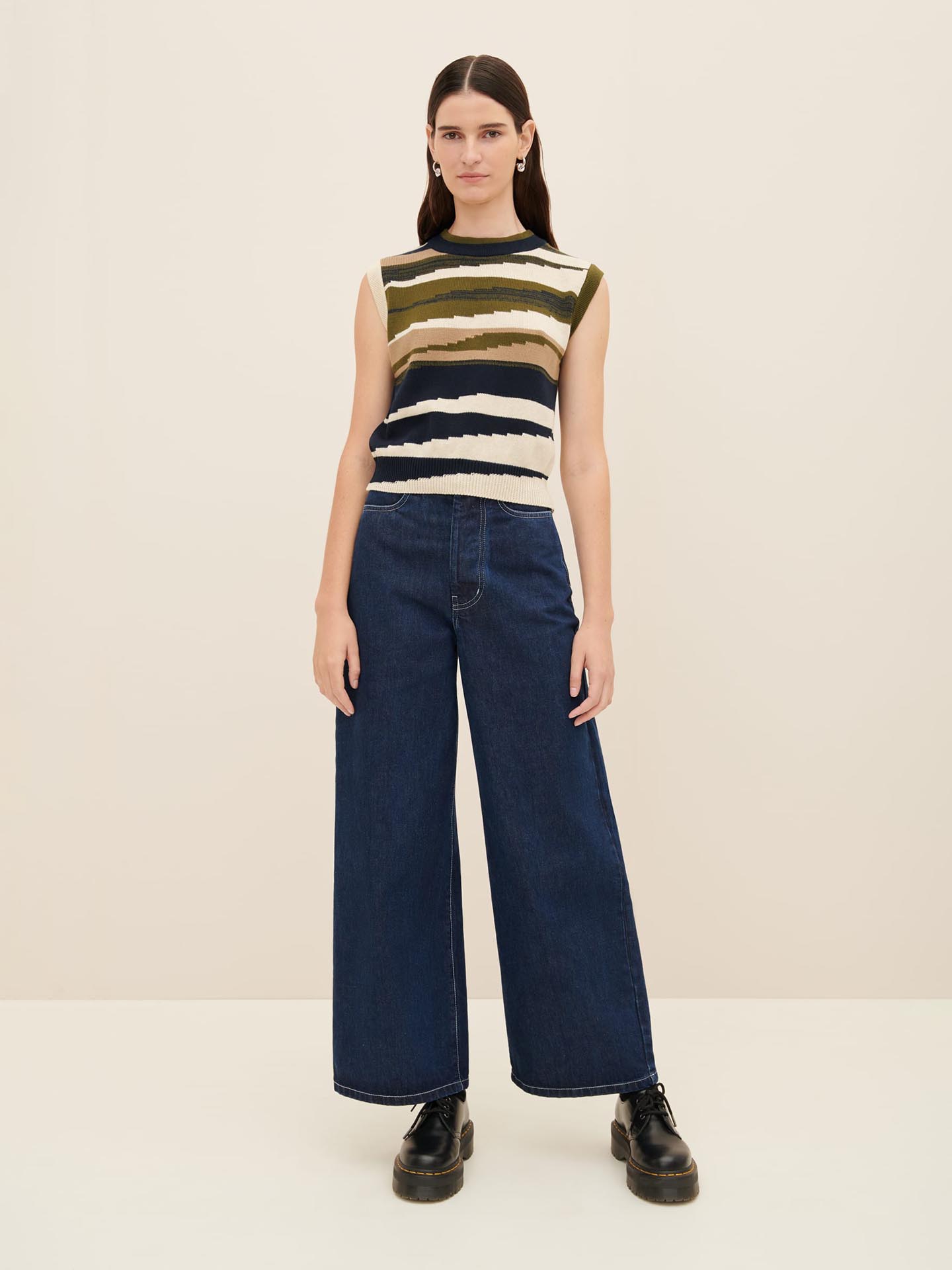 A person stands against a plain background, wearing the Kowtow Landscape Vest with horizontal stripes and wide-leg denim pants paired with black shoes, showcasing the standard fit according to the Kowtow size guide.