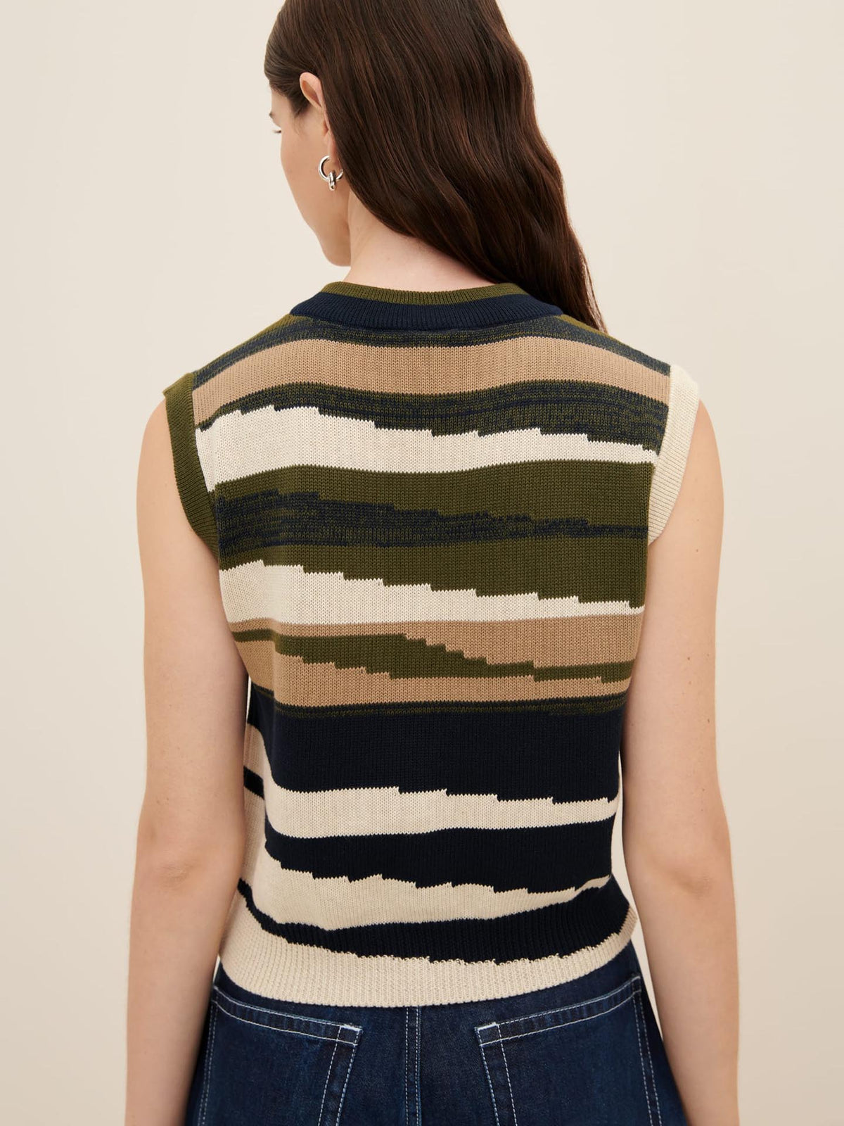 A person with long hair is shown from the back, wearing the Kowtow Landscape Vest, a sleeveless knit top featuring stripes in shades of green, beige, and black. The vest embodies the standard fit as referenced in the Kowtow size guide.