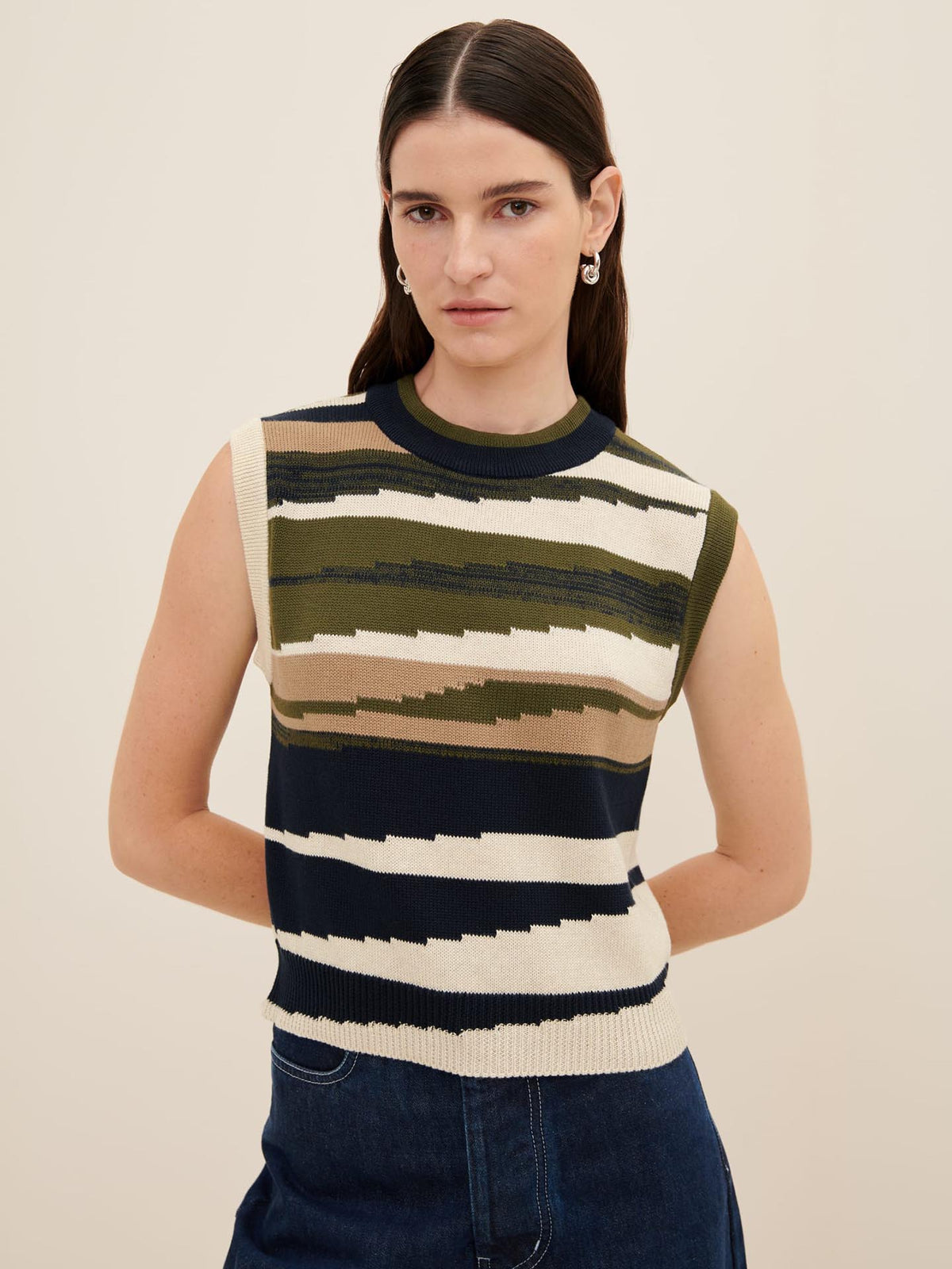 A person with long dark hair is wearing the Landscape Vest by Kowtow, a sleeveless, standard-fit sweater featuring stripes in shades of green, brown, white, and black. They have paired it with a dark denim skirt and are standing against a plain background. For accurate sizing information, refer to the Kowtow size guide.