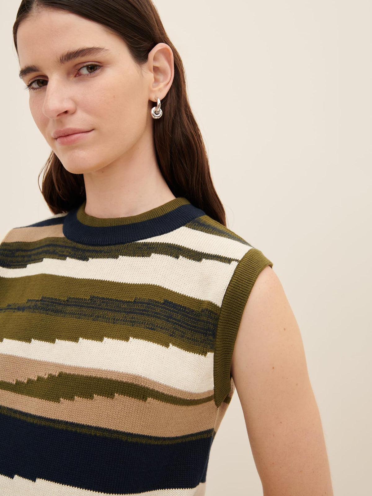 A person with long dark hair and a neutral expression wears the Kowtow Landscape Vest, featuring stripes and a sleeveless design, complemented by an earring in an abstract shape, exemplifying the perfect standard fit as outlined in the Kowtow size guide.