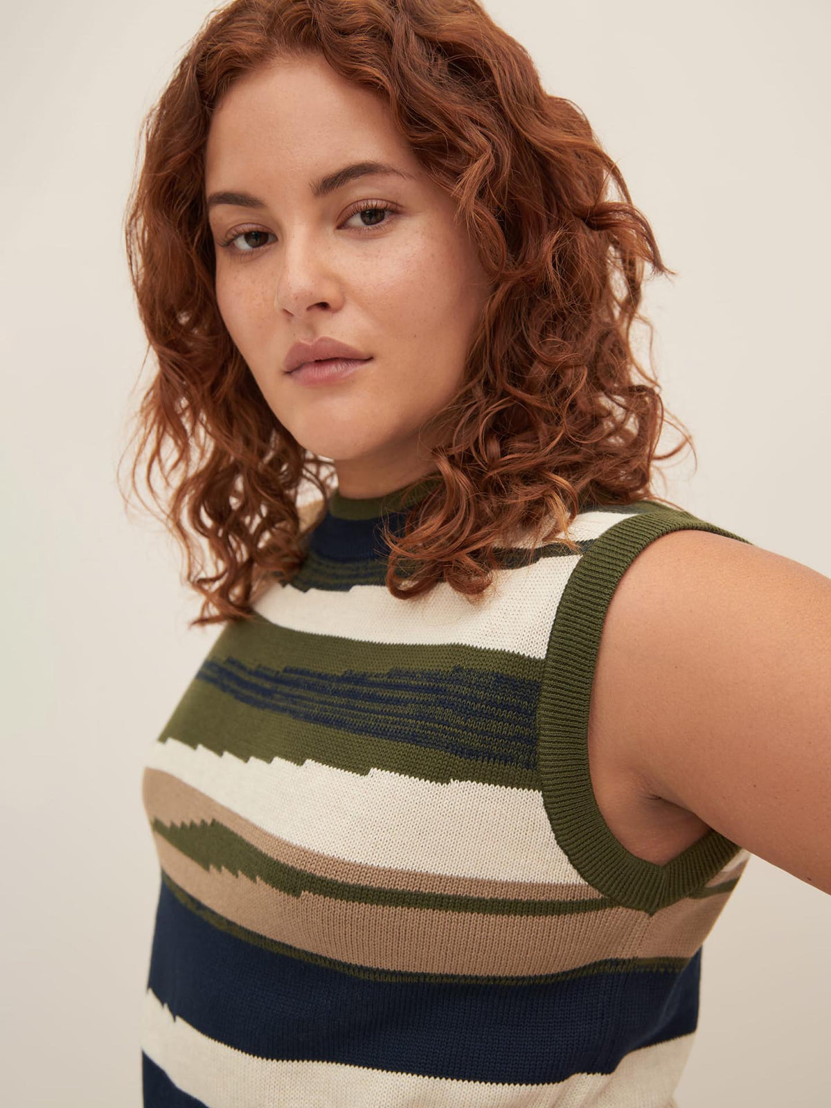 A person with curly red hair, wearing the Kowtow Landscape Vest, stands against a plain background. The sleeveless top features multicolored stripes and effortlessly embodies the standard fit showcased in the Kowtow size guide as they look slightly to the side.