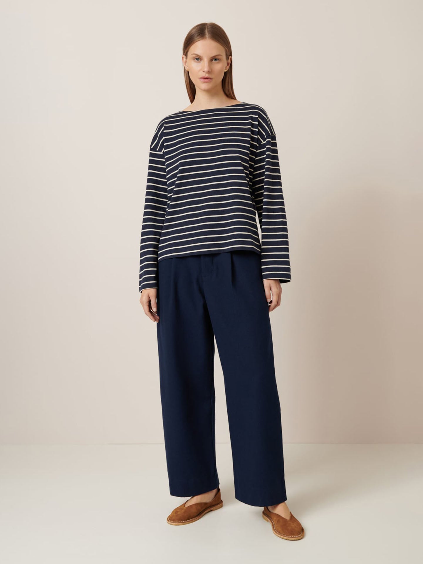 Against a plain background, a person stands wearing the Leo Top – Sailor Stripe by Kowtow paired with relaxed-fit navy wide-leg pants and brown shoes.