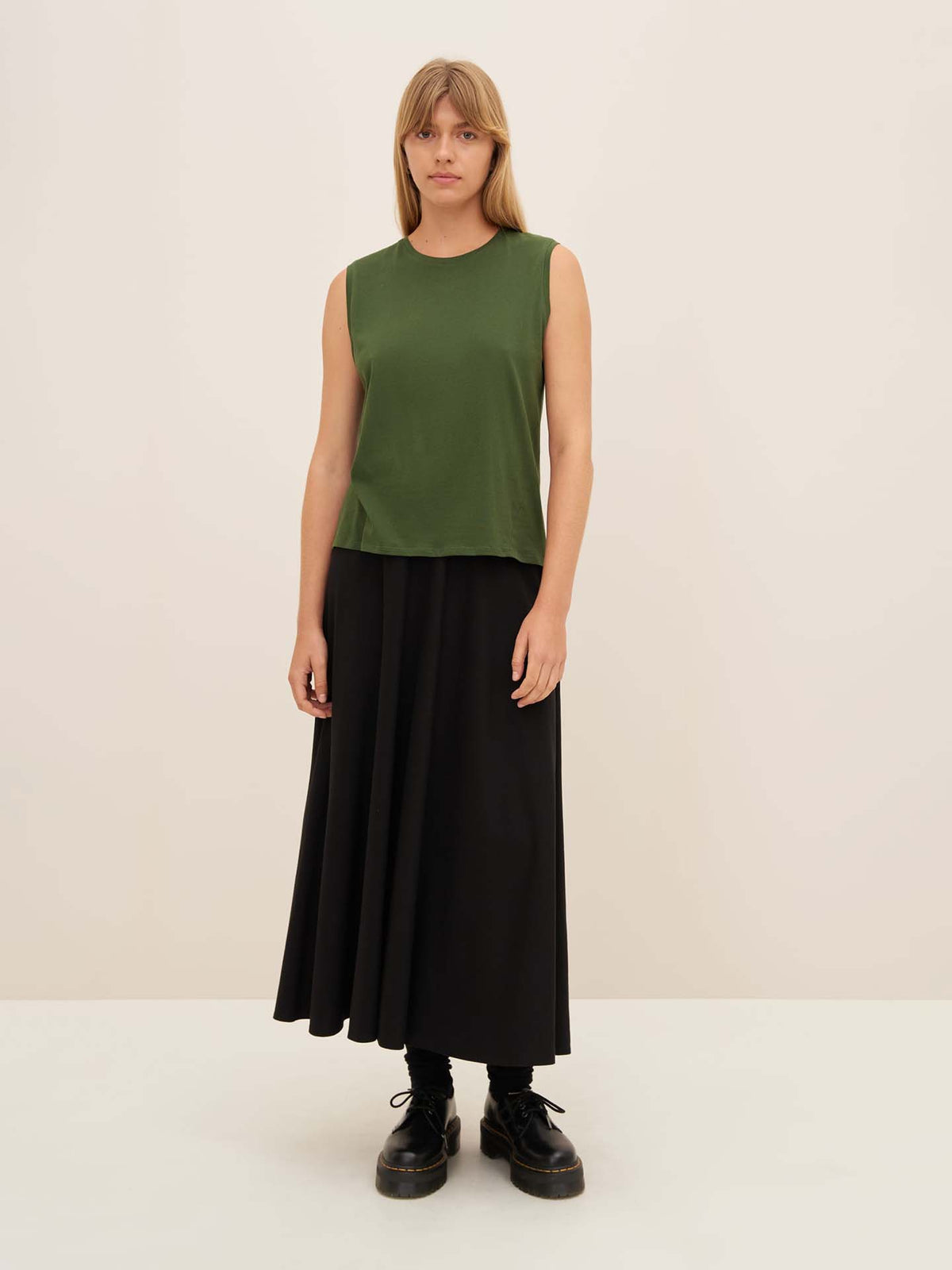 A person wearing the Kowtow Light Singlet in Forest, a green sleeveless top, paired with a black maxi skirt, both featuring a relaxed fit and true to size, stands against a plain background in black shoes.