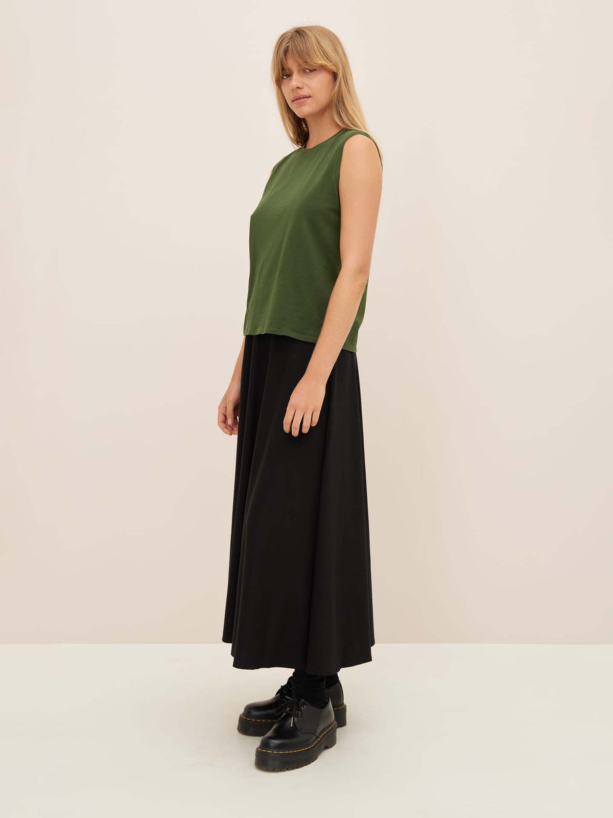 A person with long hair is wearing the Kowtow Light Singlet in Forest, a green sleeveless top designed for a relaxed fit, paired with a black long skirt and matching boots, standing against a plain background.