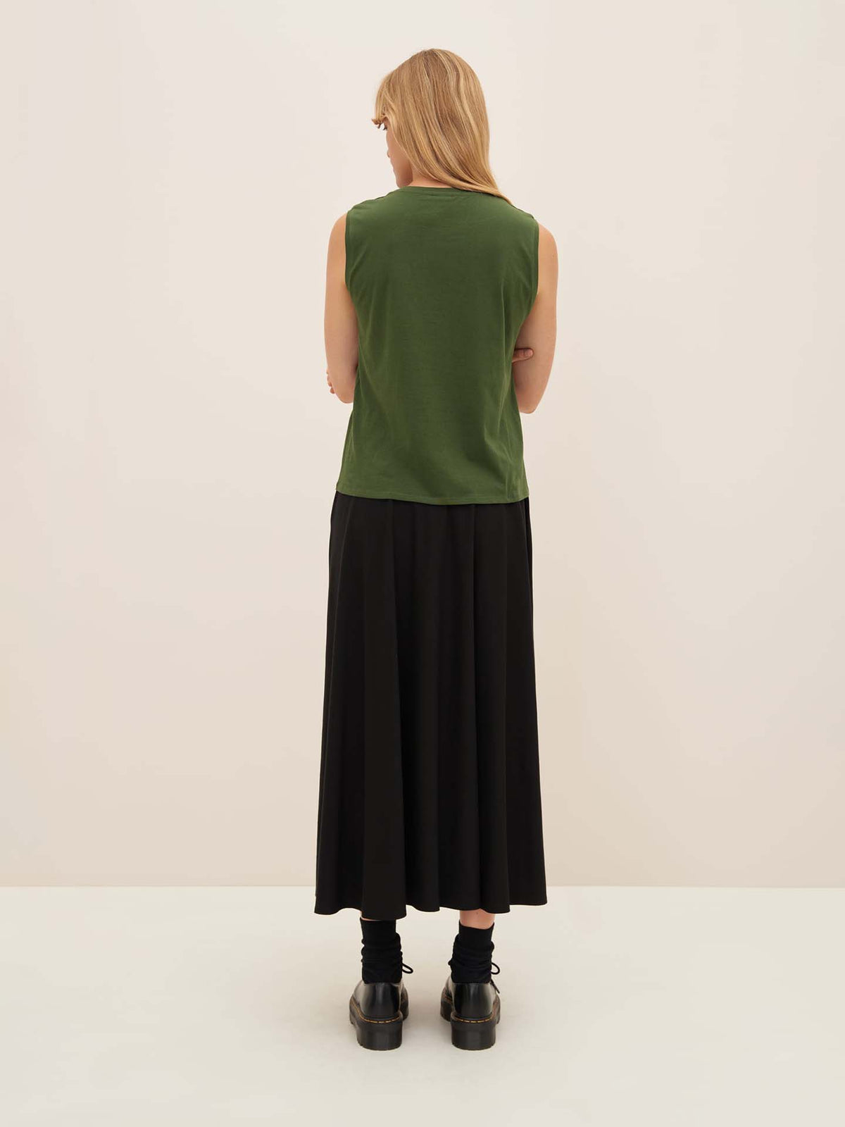 A person with long hair, wearing the Kowtow size XS Light Singlet in Forest and a black relaxed-fit skirt, stands against a plain background, black shoes completing the look.