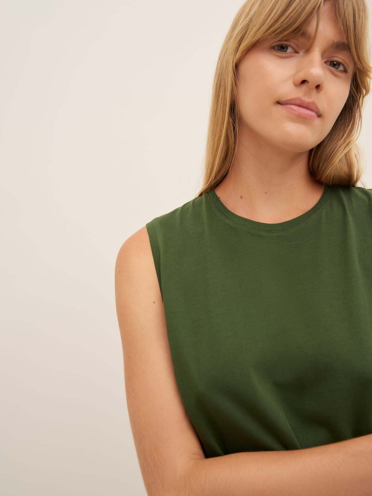 A person with long hair is wearing Kowtow’s Light Singlet in Forest, designed in a relaxed fit, while standing against a plain, neutral background.