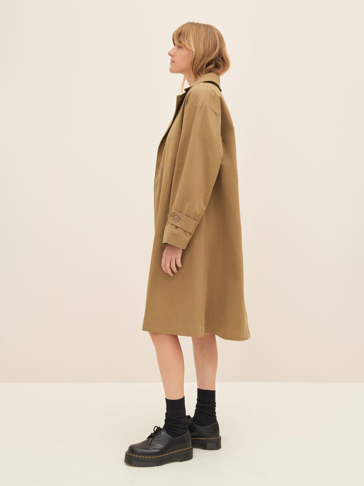 A person wearing the Kowtow Mile Trench in Bronze, black socks, and black shoes stands sideways against a plain background.