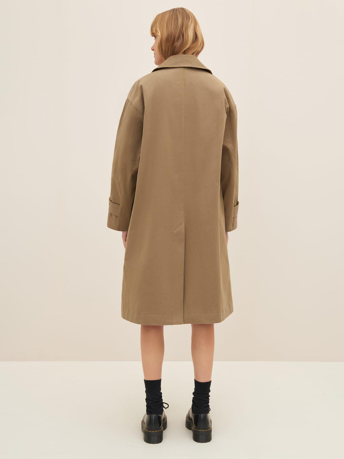 Person wearing an oversized fit knee-length bronze Mile Trench coat by Kowtow, a black skirt, and black shoes, standing with their back to the camera.