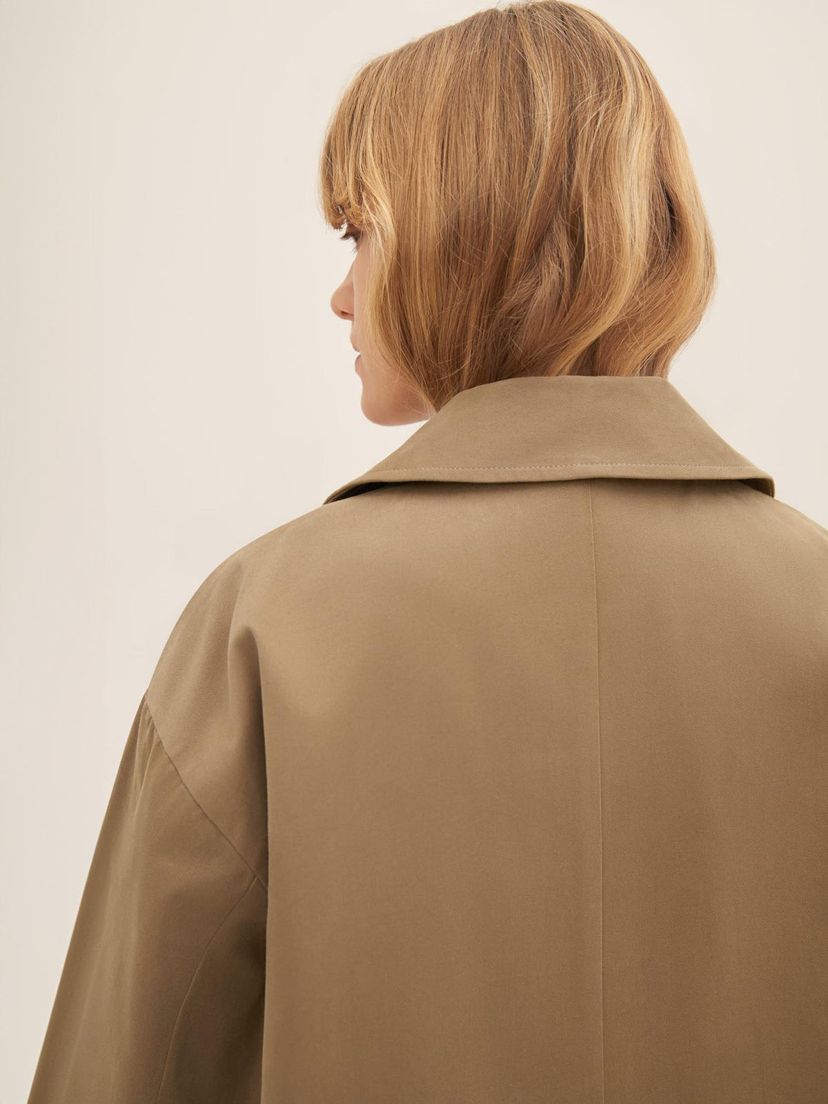 A person with shoulder-length hair is seen from behind, wearing the Mile Trench – Bronze by Kowtow, featuring a wide collar and an oversized fit.