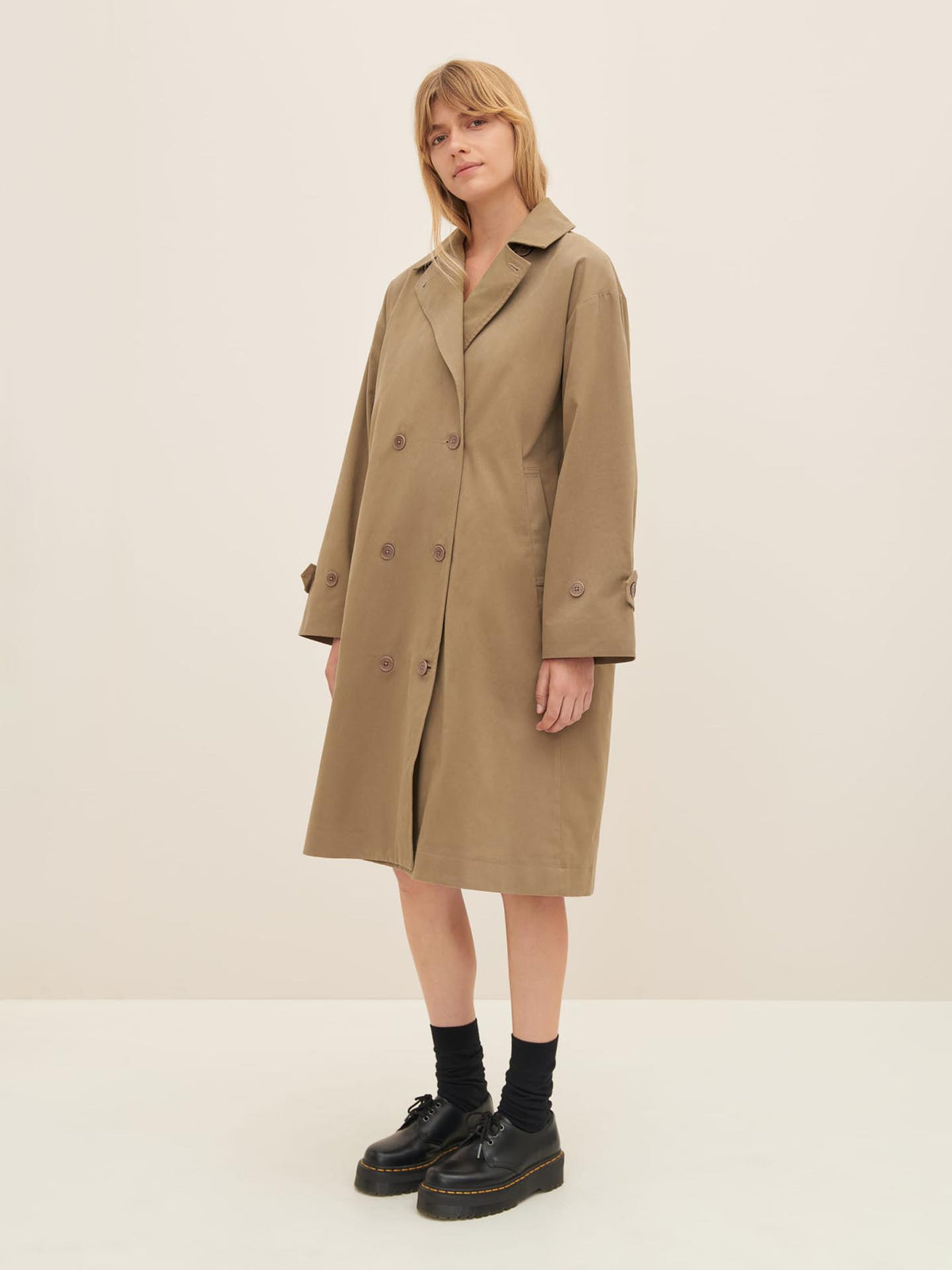 A person stands against a plain background wearing the Mile Trench in Bronze by Kowtow, showcasing the oversized fit perfectly, complemented by black socks and black shoes.