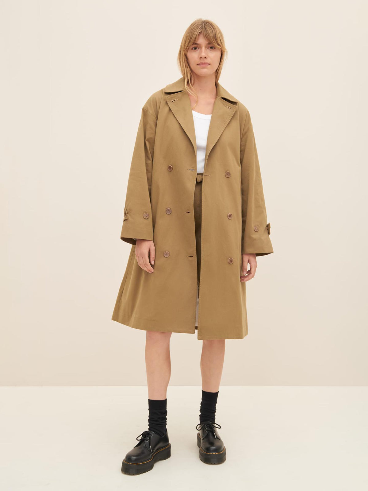 A person stands against a plain background, wearing the Mile Trench in bronze with an oversized fit over a white top and black shoes, showcasing the relaxed sophistication of Kowtow&#39;s latest collection.