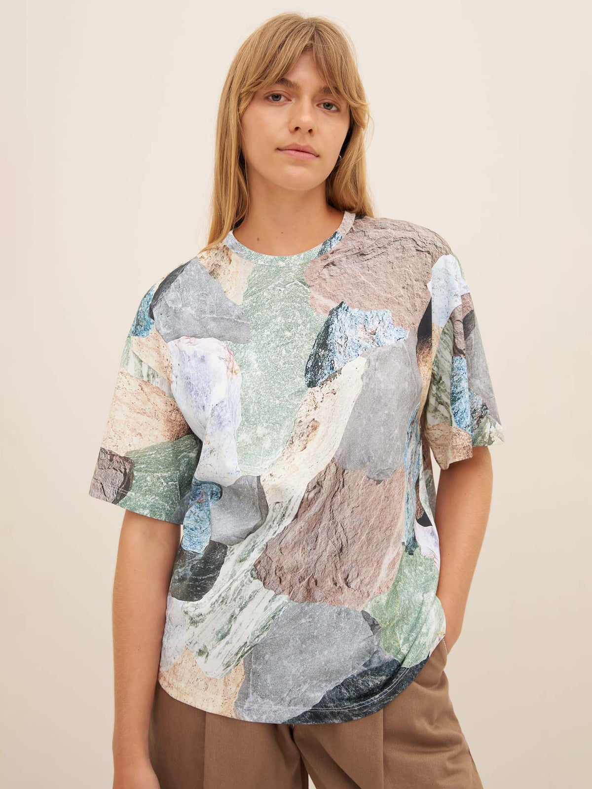 A person wearing Kowtow&#39;s Mineral Tee, featuring a colorful abstract pattern and an oversized fit, along with brown pants, stands against a plain background.